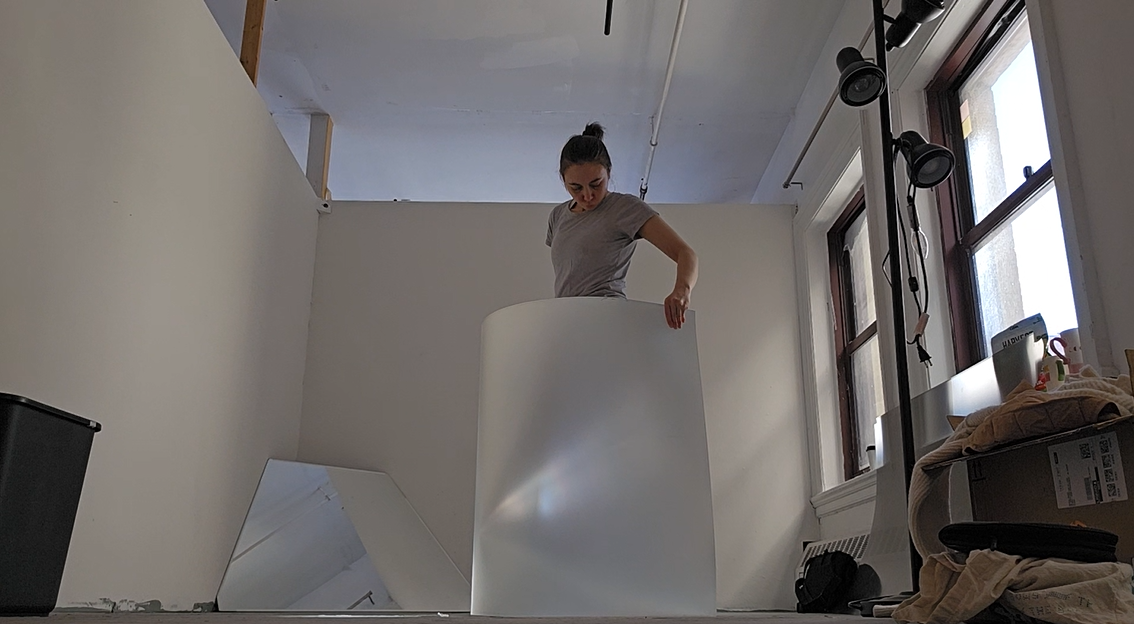 A woman stands in the middle of a sunlight room in waning light. She wears a t-shirt and has brown hair in a bun. She holds a giant fresnel lens around her body, looking down to see just reflected light and the disappearance of her body. 