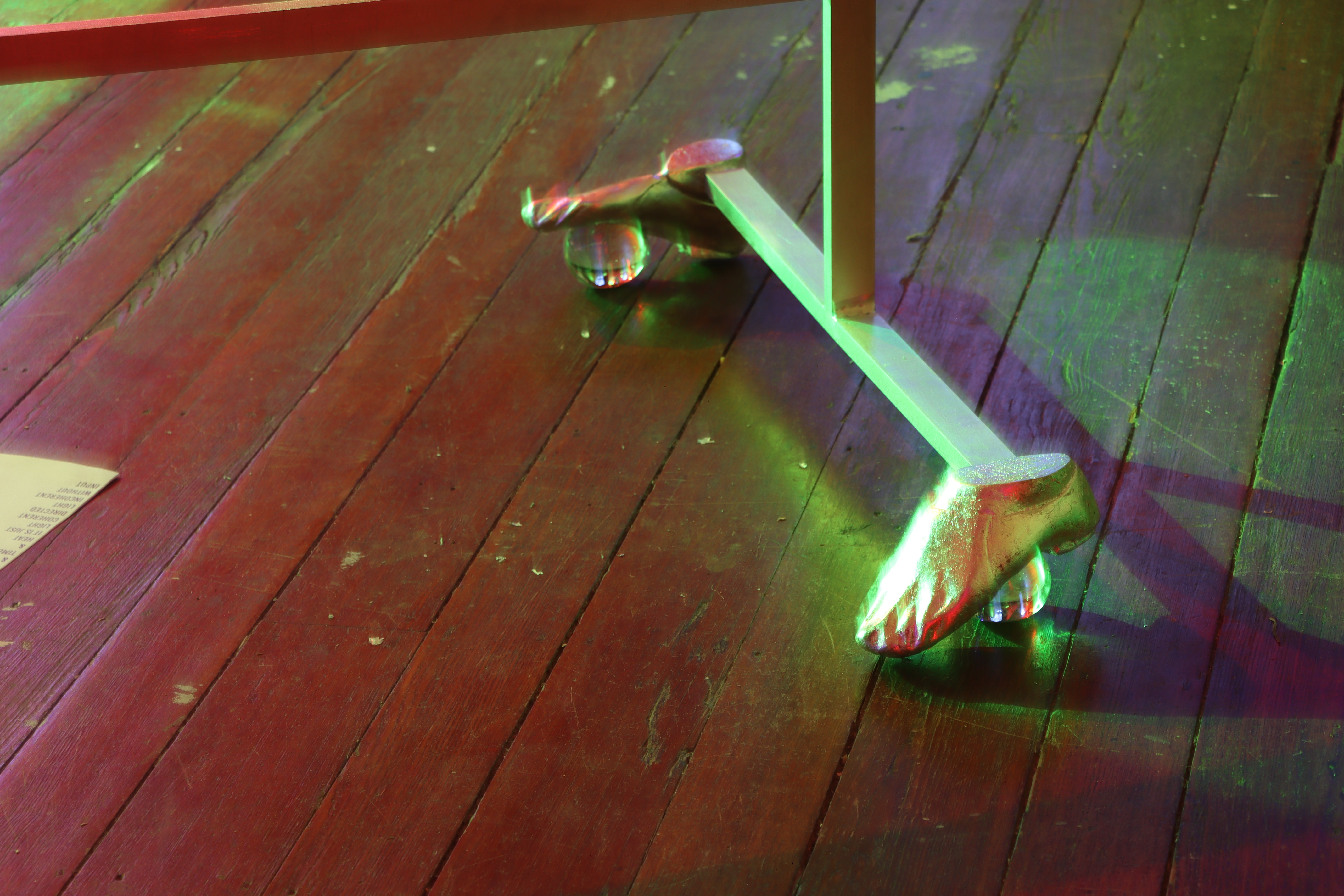 Close up of a silver sculpture base, whose feet are aluminum feet in jazz shoes, over solid glass balls. The light is green from one direction, and red from another, fuzzy and a little out of focus. 
