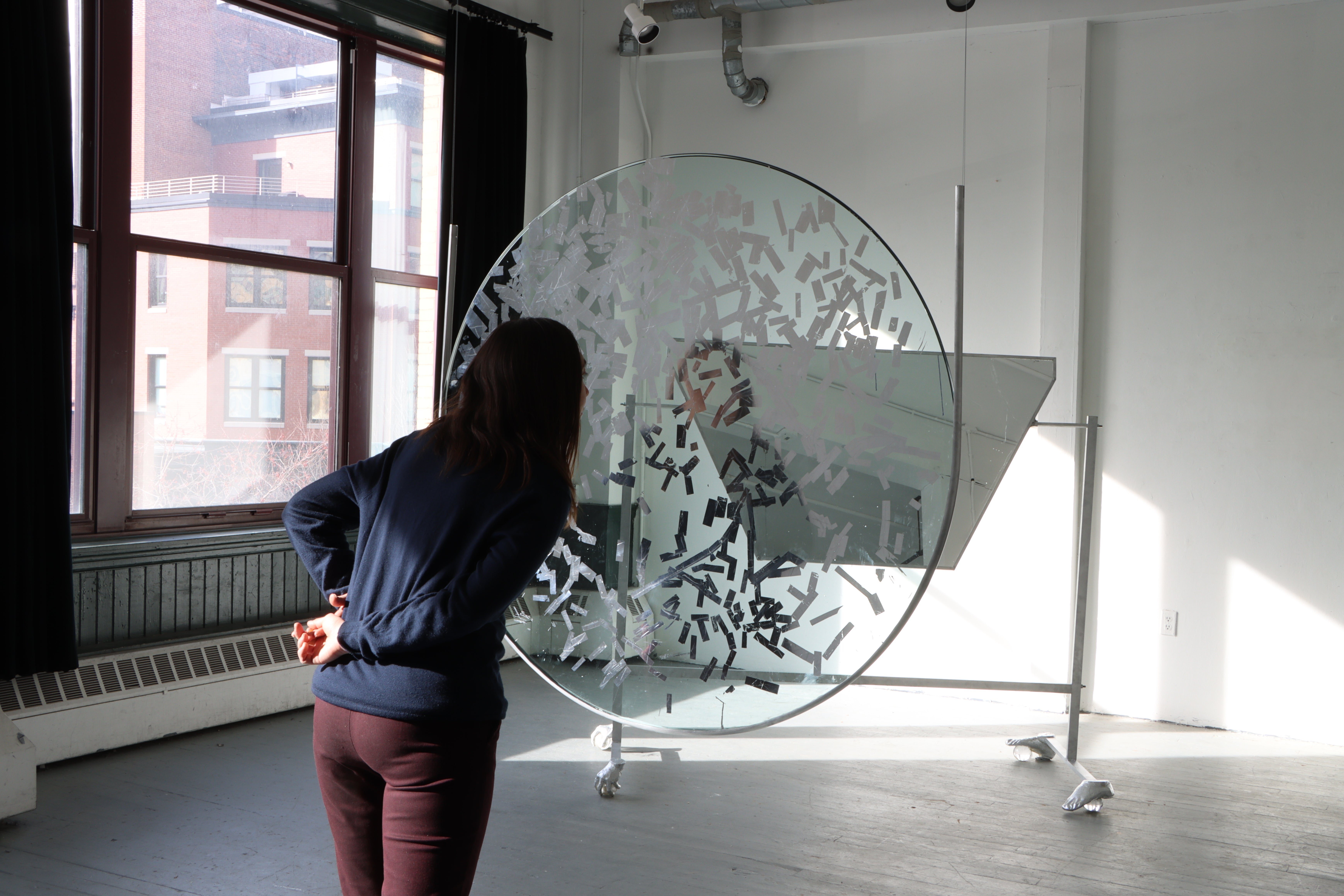 4ft diameter piece of glass hands supsended in a room, with part of its surfaced gilded silver. A person in motion passes behind it. Behind them is large piece of mirror angled on a aluminum stand, with feet made of hands and feet on crystal balls.