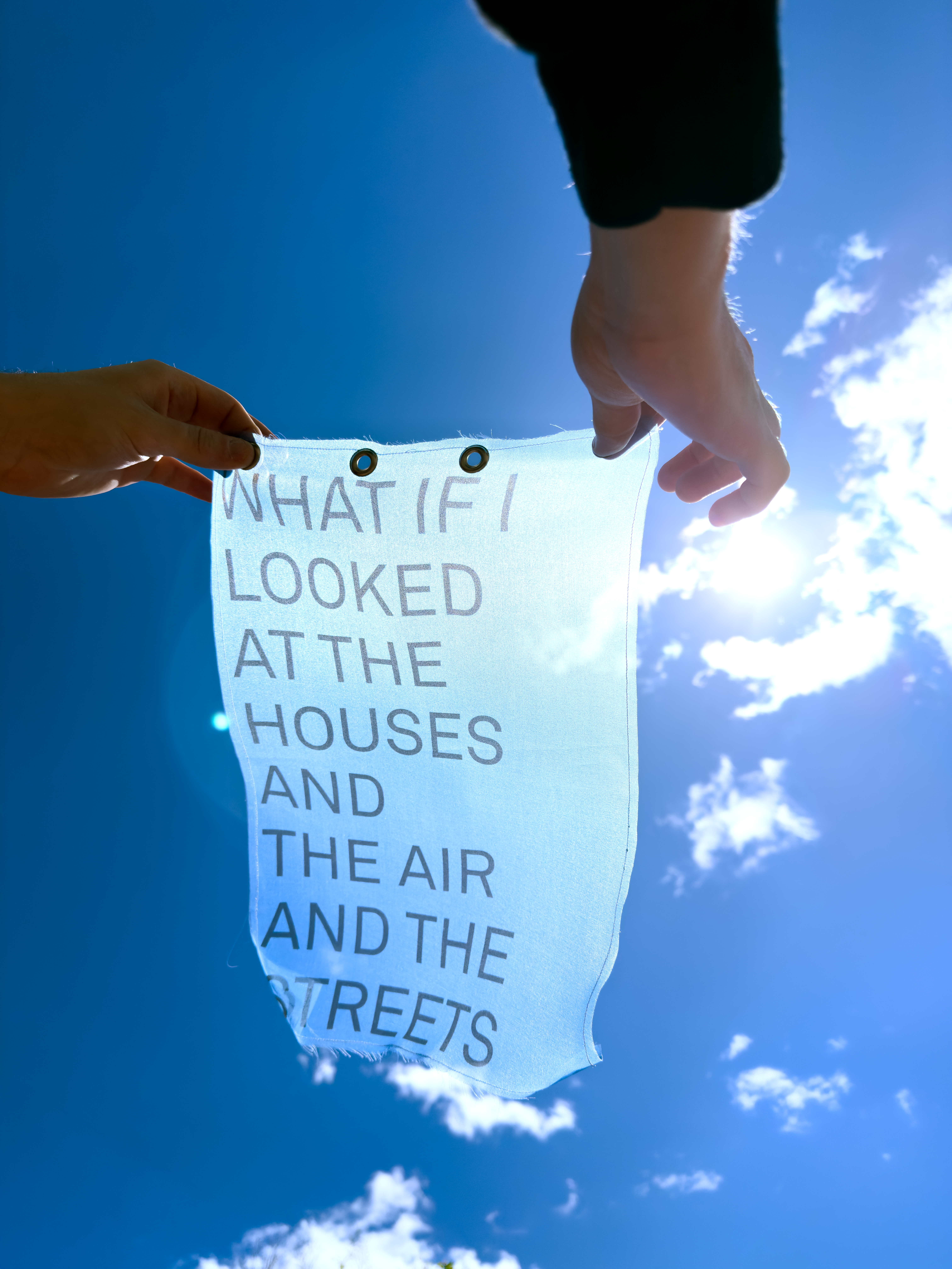 Someone holding a blue fabric print up to the sky that reads "what if I looked at the houses, and the air, and the streets