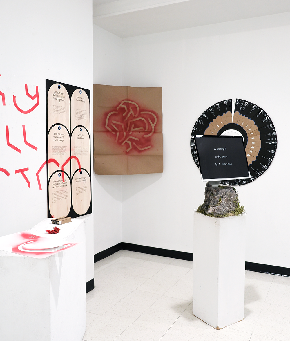 An installation critical of AI, showing an iMac within a sculpted rock on a pedestal, various red, black, and tan typographic objects on walls