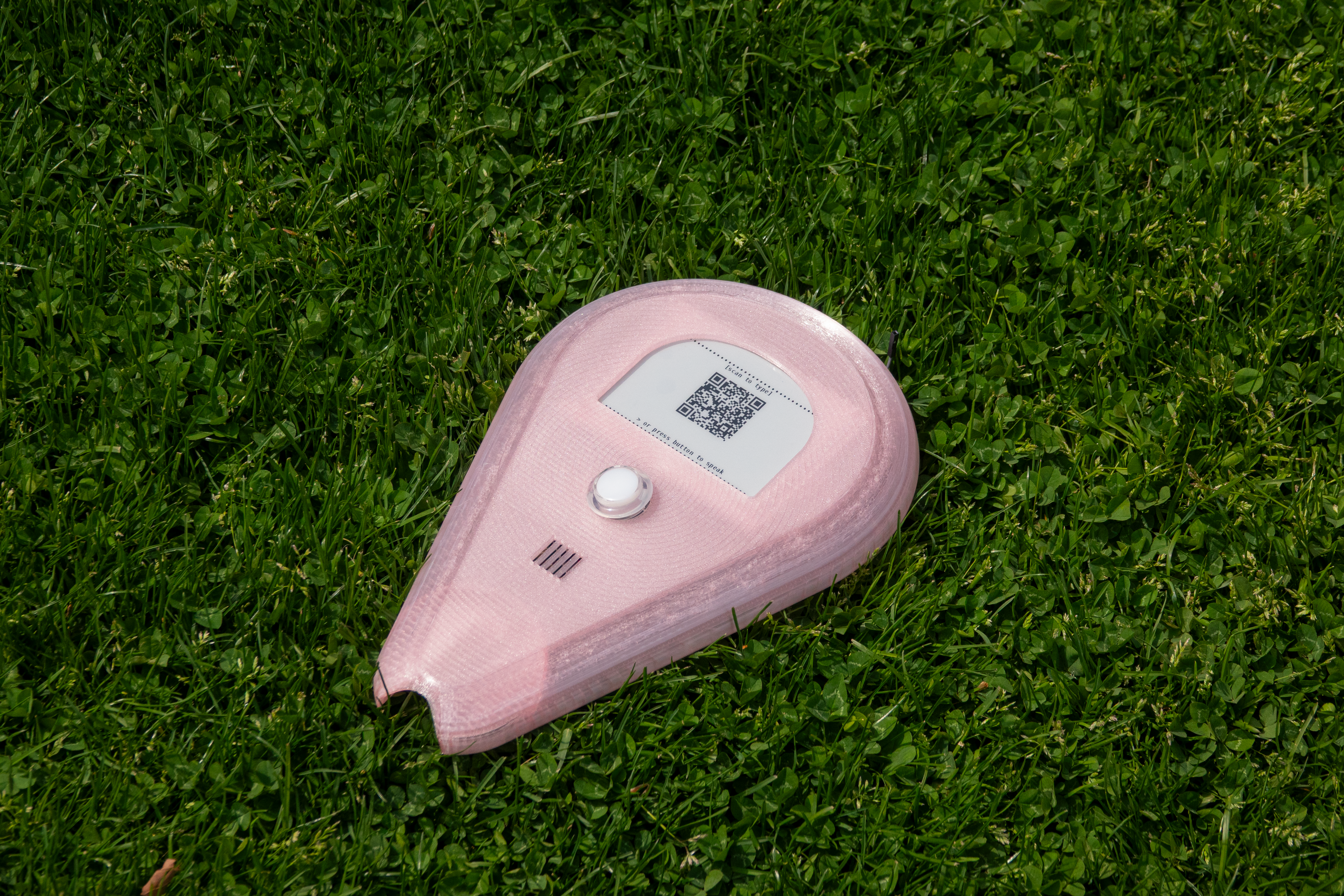 Pink teardrop-shaped interactive device resting on grass, featuring a button, speaker, and QR code displayed on a screen.