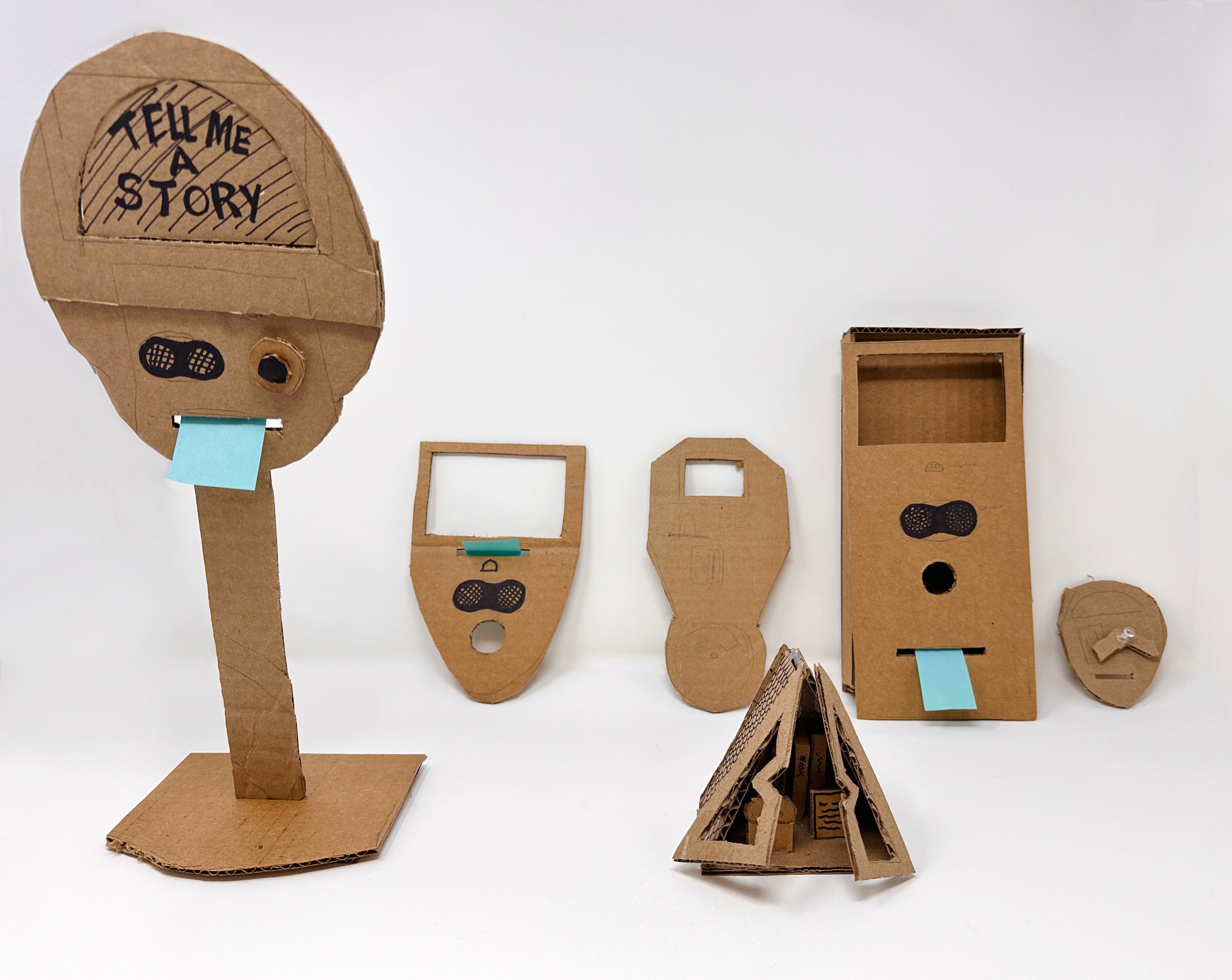 Six handmade cardboard prototypes of interactive objects, each with speaker grilles, a button hole, and a blue sticky-note "receipt" slot. The tallest reads "Tell Me A Story."