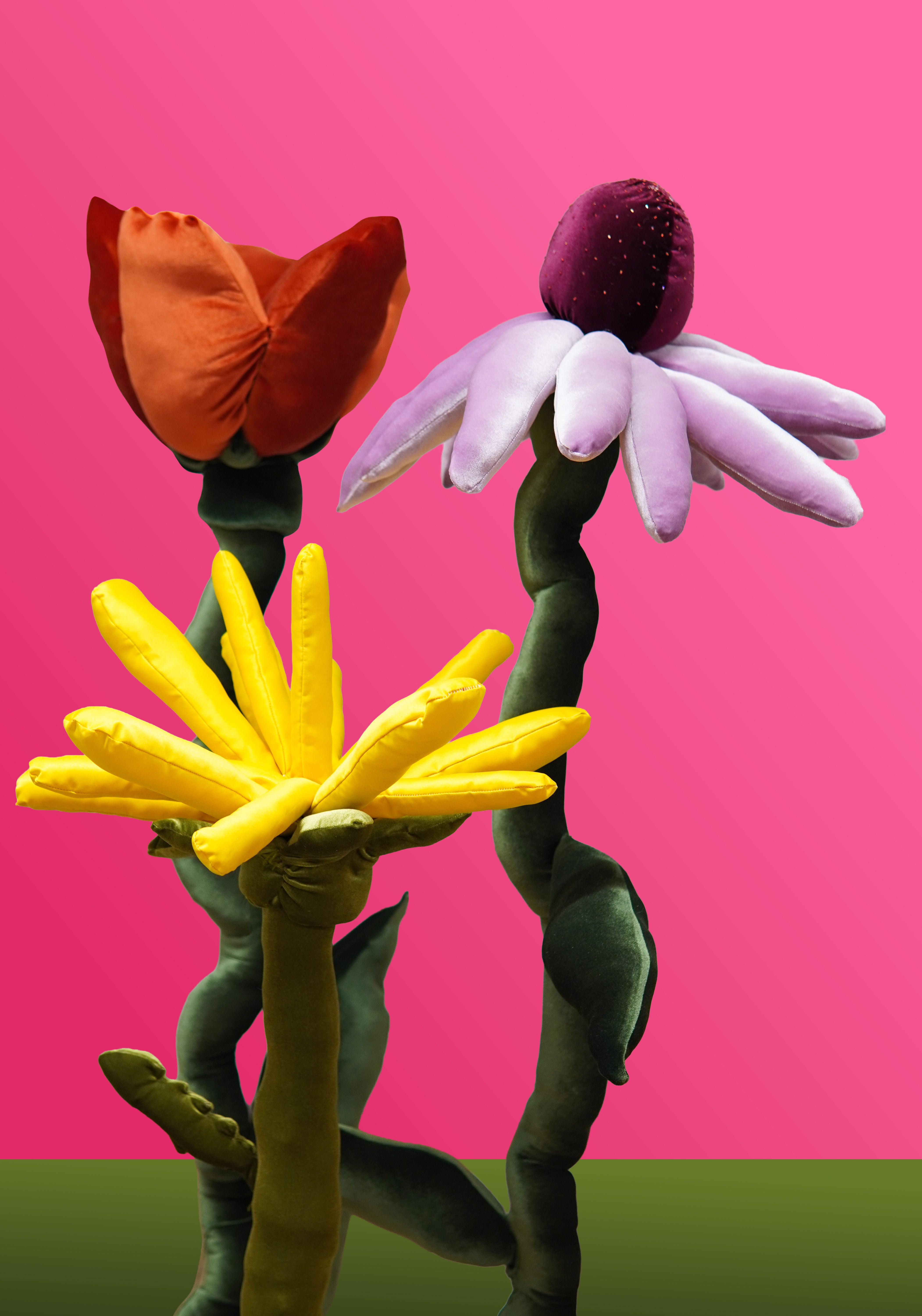 Oversized soft sculptures of three colorful flowers with velvet petals and twisting green stems, displayed against a bright pink background.