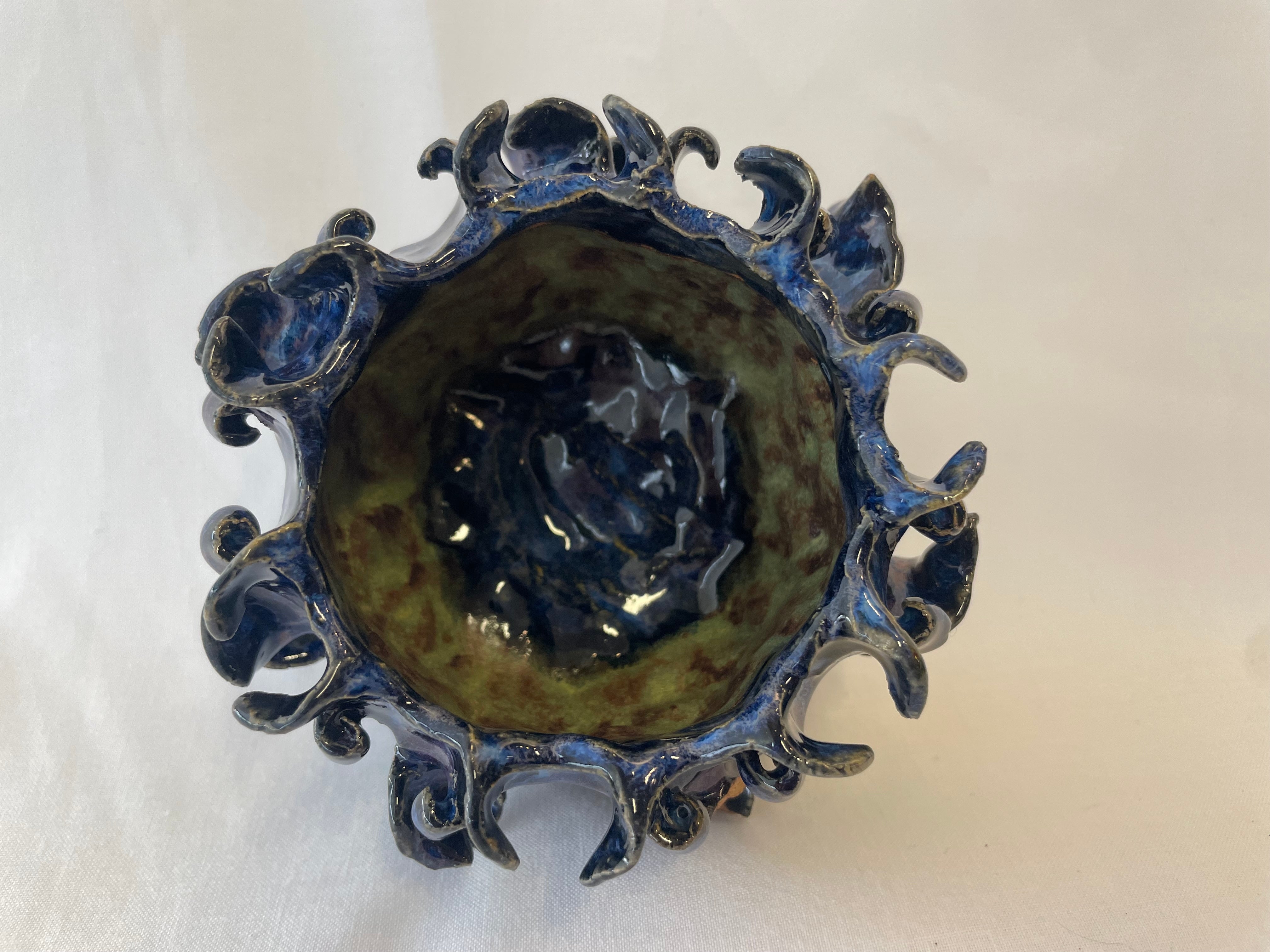 Ceramic bowl with leaf-like shapes surrounding the outside going in different pattern directions. Glaze colors are a  dark navy blue primarily with a green stripe on the inside.