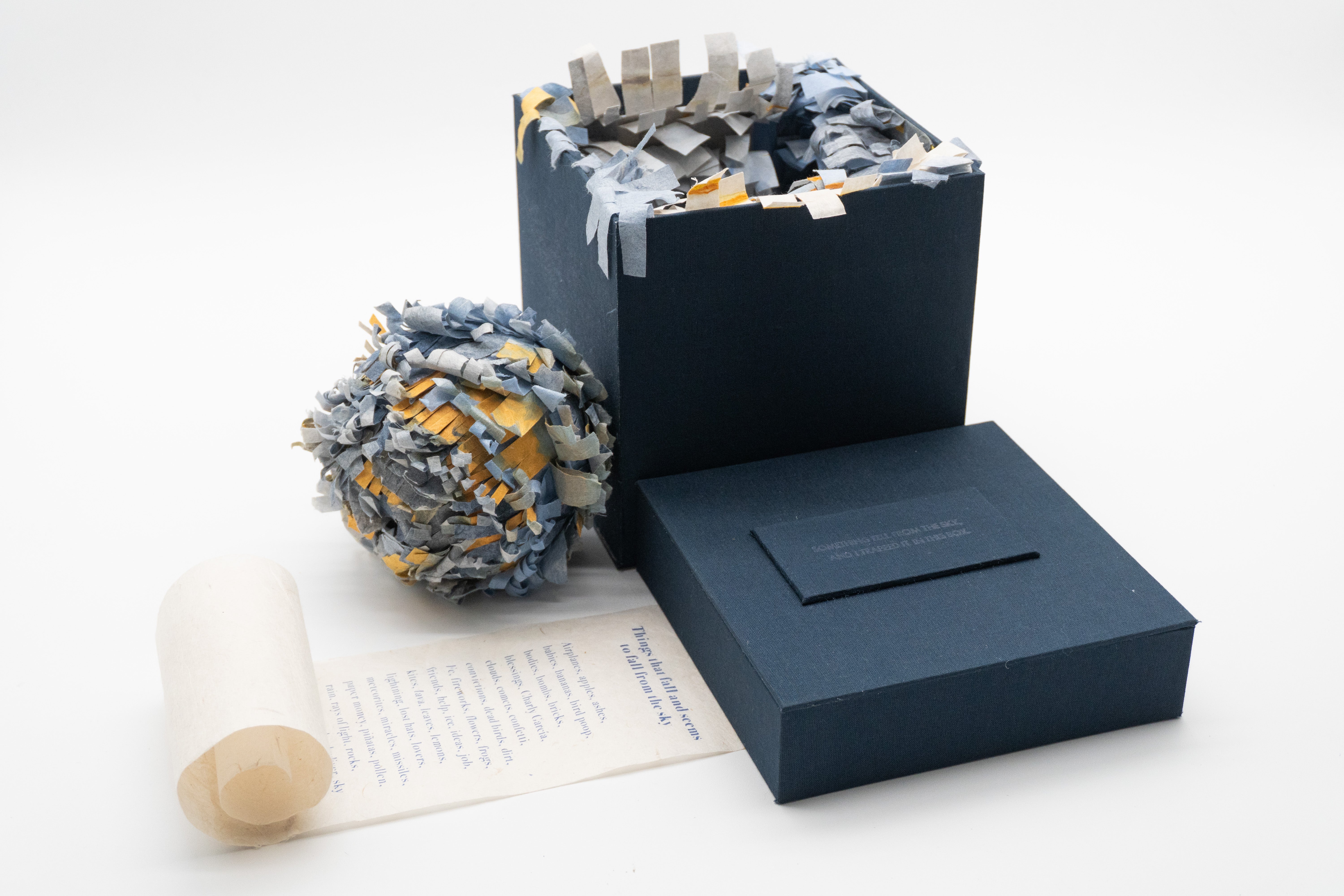 A small installation shaped like a book consists of a dark blue square box, a rolled handmade paper scroll, and a spherical handmade piñata covered in layered blue, gray, white, and yellow paper fragments.
