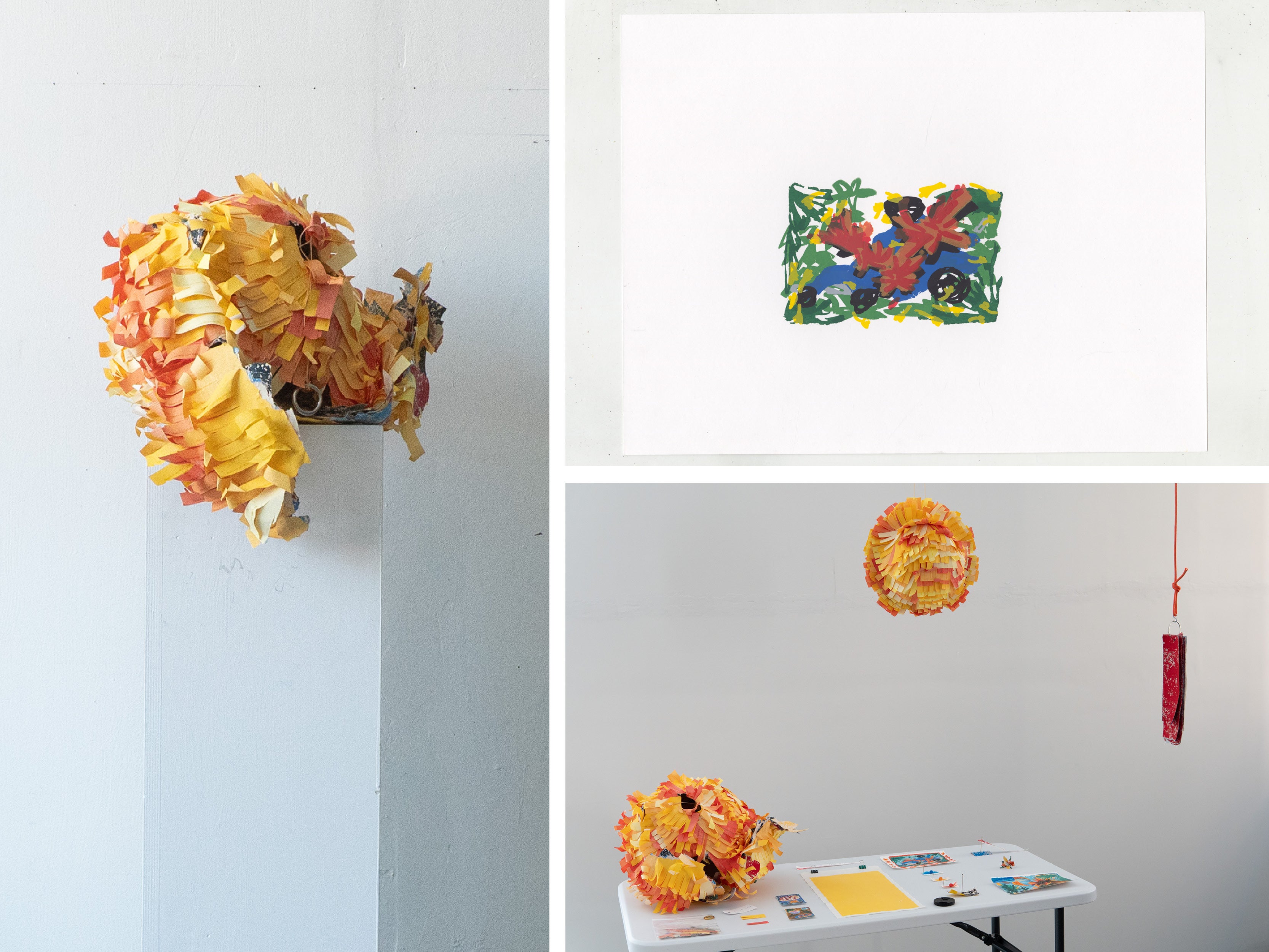 An installation features large piñatas made from layered strips of yellow, orange, and pink paper. One sculpture sits on a white pedestal, while another hangs from the ceiling above a white table displaying small printed images