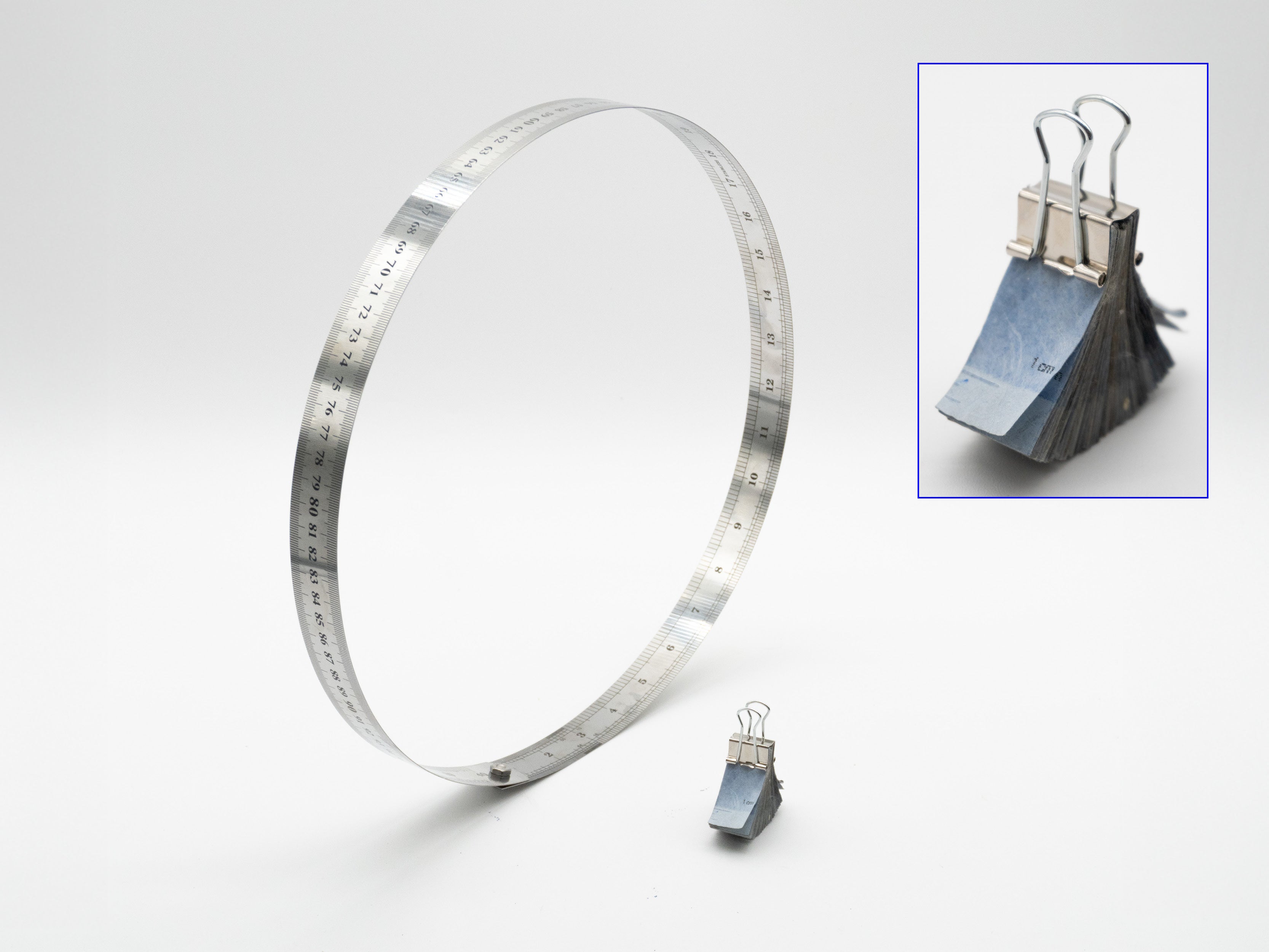 A metallic ruler forms a large standing loop against a white background, bending away from its flat shape and held together with a small screw. Beside it sits a small stack of blue intaglio-printed paper pieces