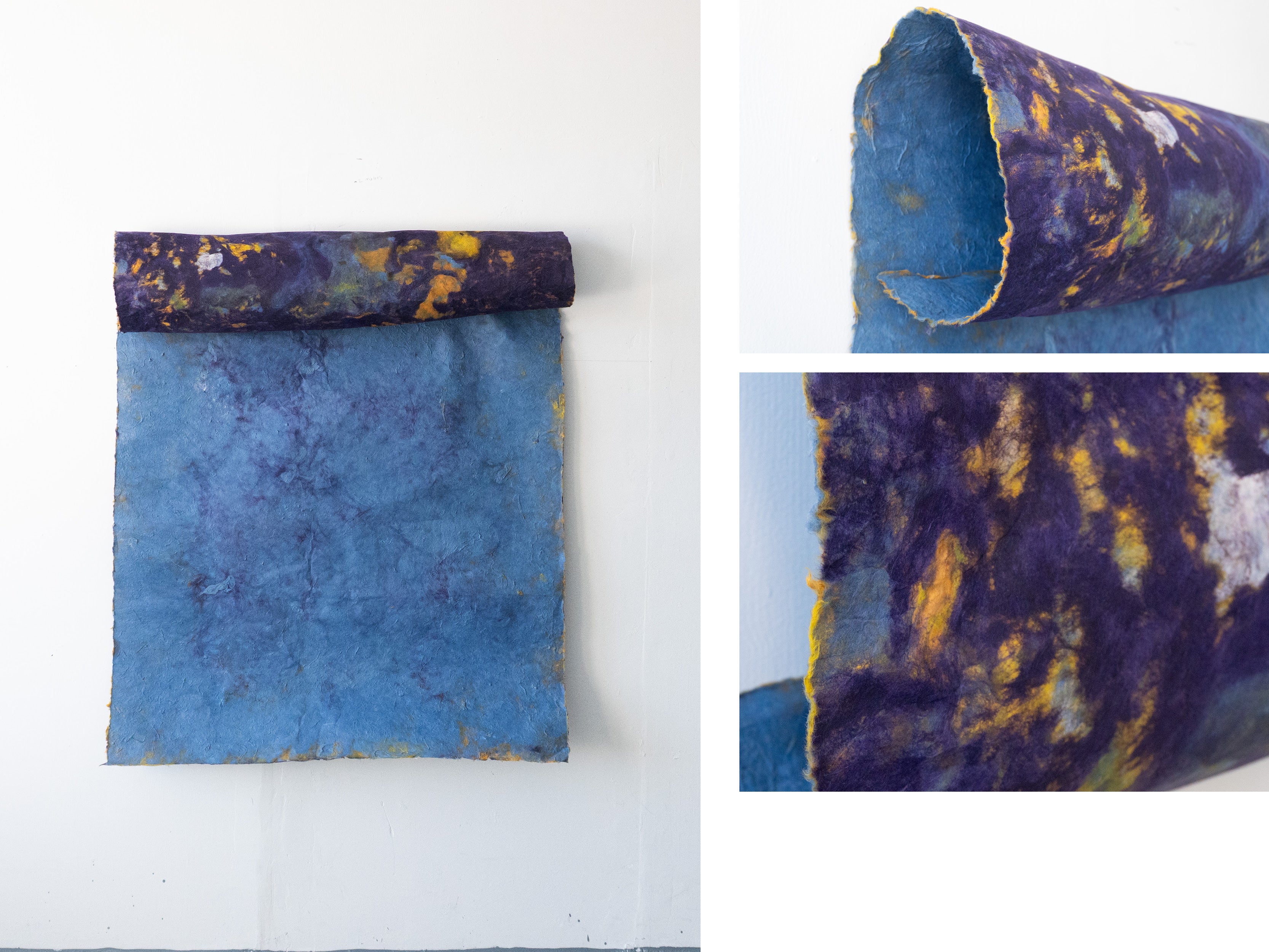 A large sheet of handmade blue paper hangs from a rolled cylindrical form attached along its top edge. The cylinder is covered in marbled purple, yellow, white, and blue textures. Close-up images show the rough edges