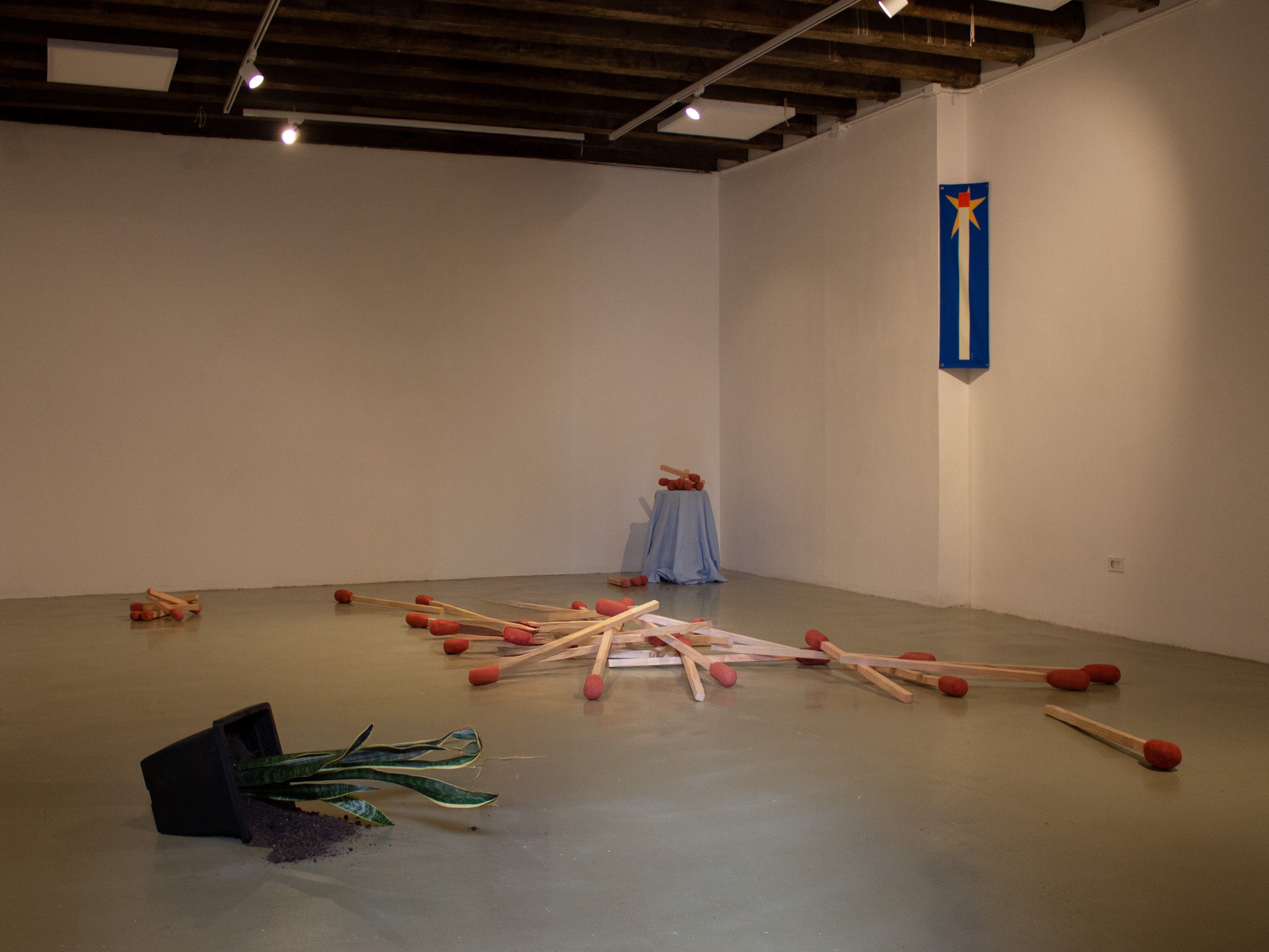 An installation fills a gallery floor with oversized wooden matches scattered in different directions. Each match has a large red tip and pale wooden shaft. In the foreground, a black flower pot lies tipped over with green paper leaves spilling out.
