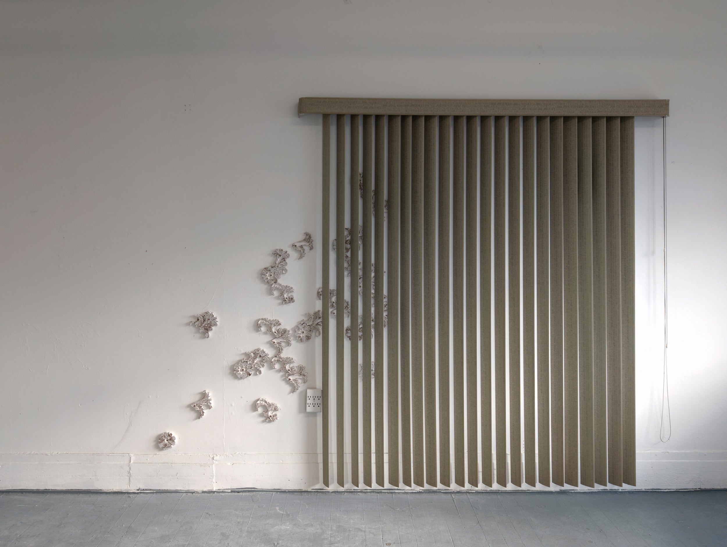 A set of vertical greenish blinds set on a white wall, a pattern of white flowers seep behind it.