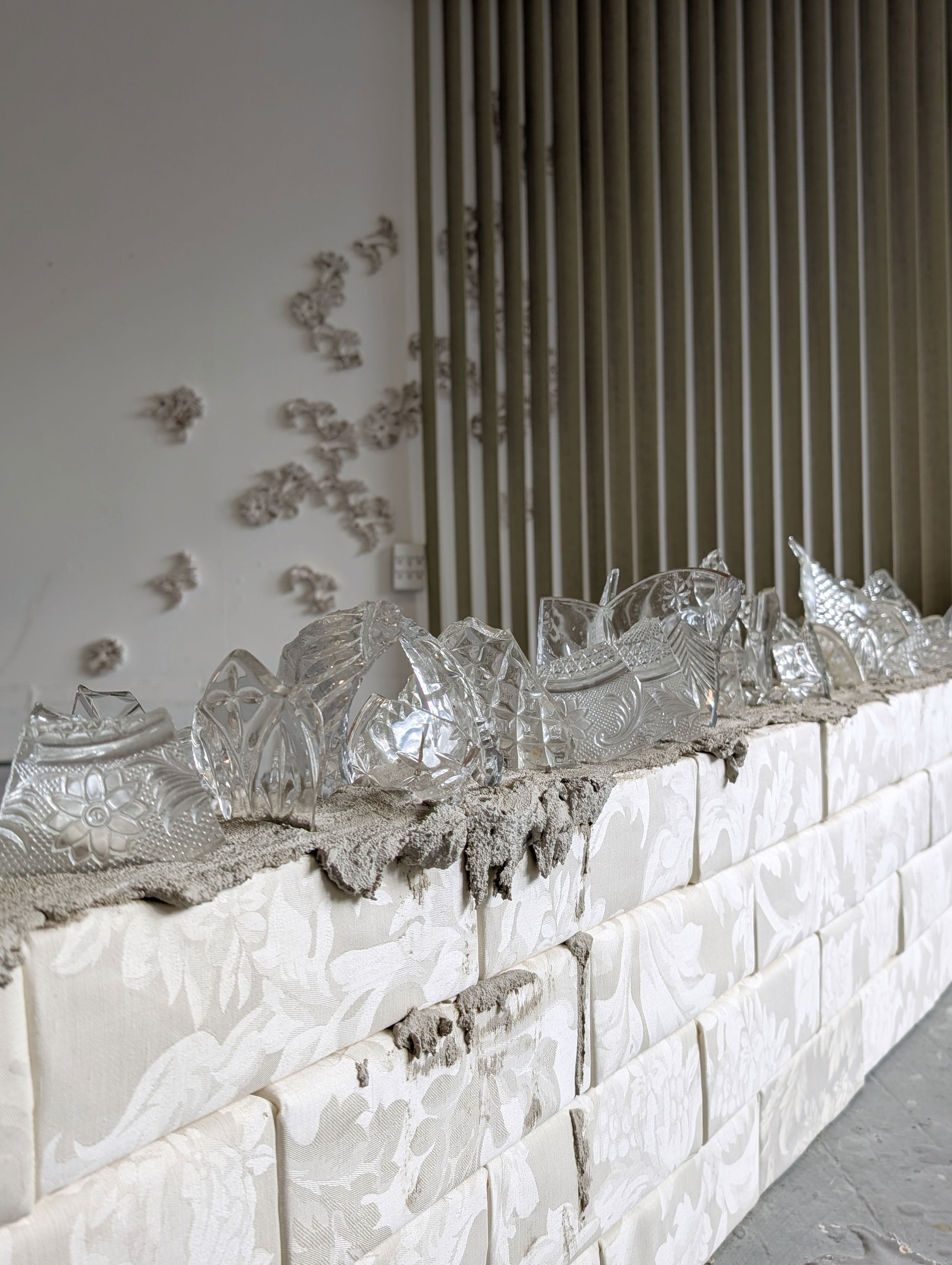 Broken antique glassware rests on a concrete bed atop a low wall built of upholstered, flower-patterned bricks.