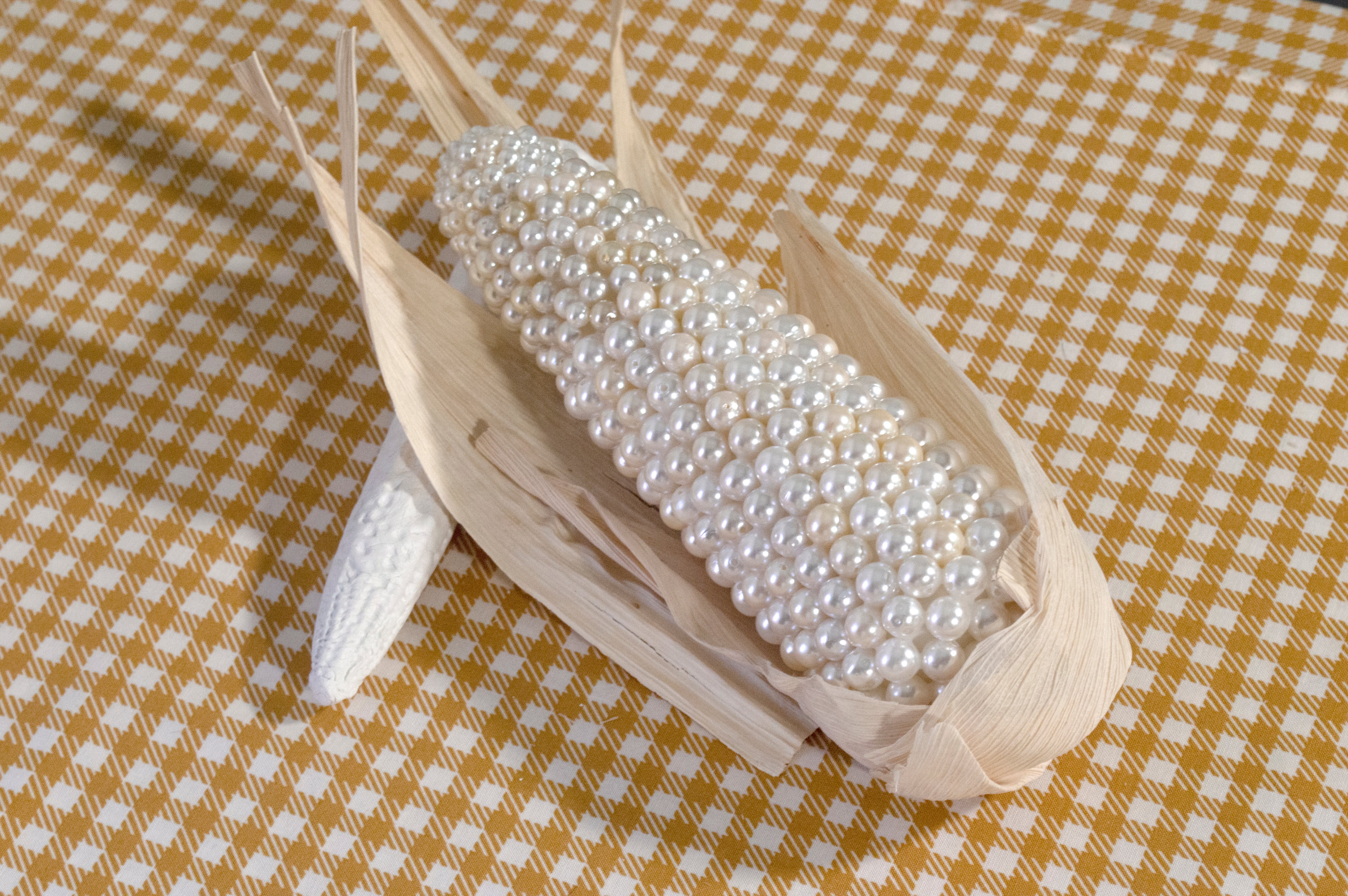 Fictional corn ear with faux pearls instead of kernels, rests on a gingham tablecloth. 