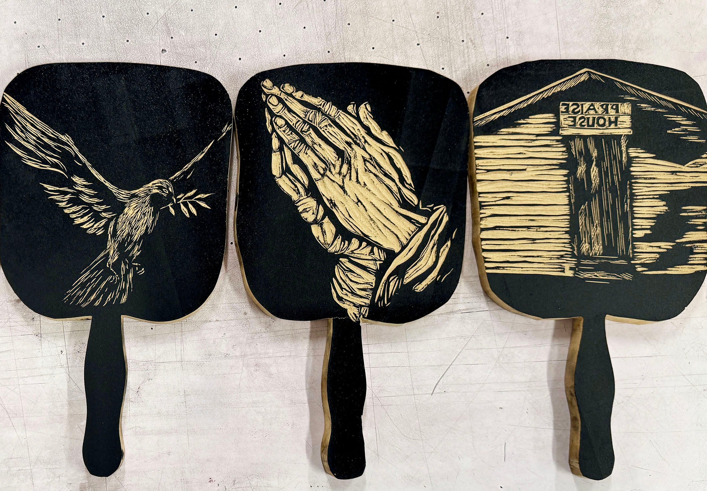 Three black and white wooden fans featuring Christian symbols: a dove with an olive branch, praying hands, and a miniature church that reads praise house. 