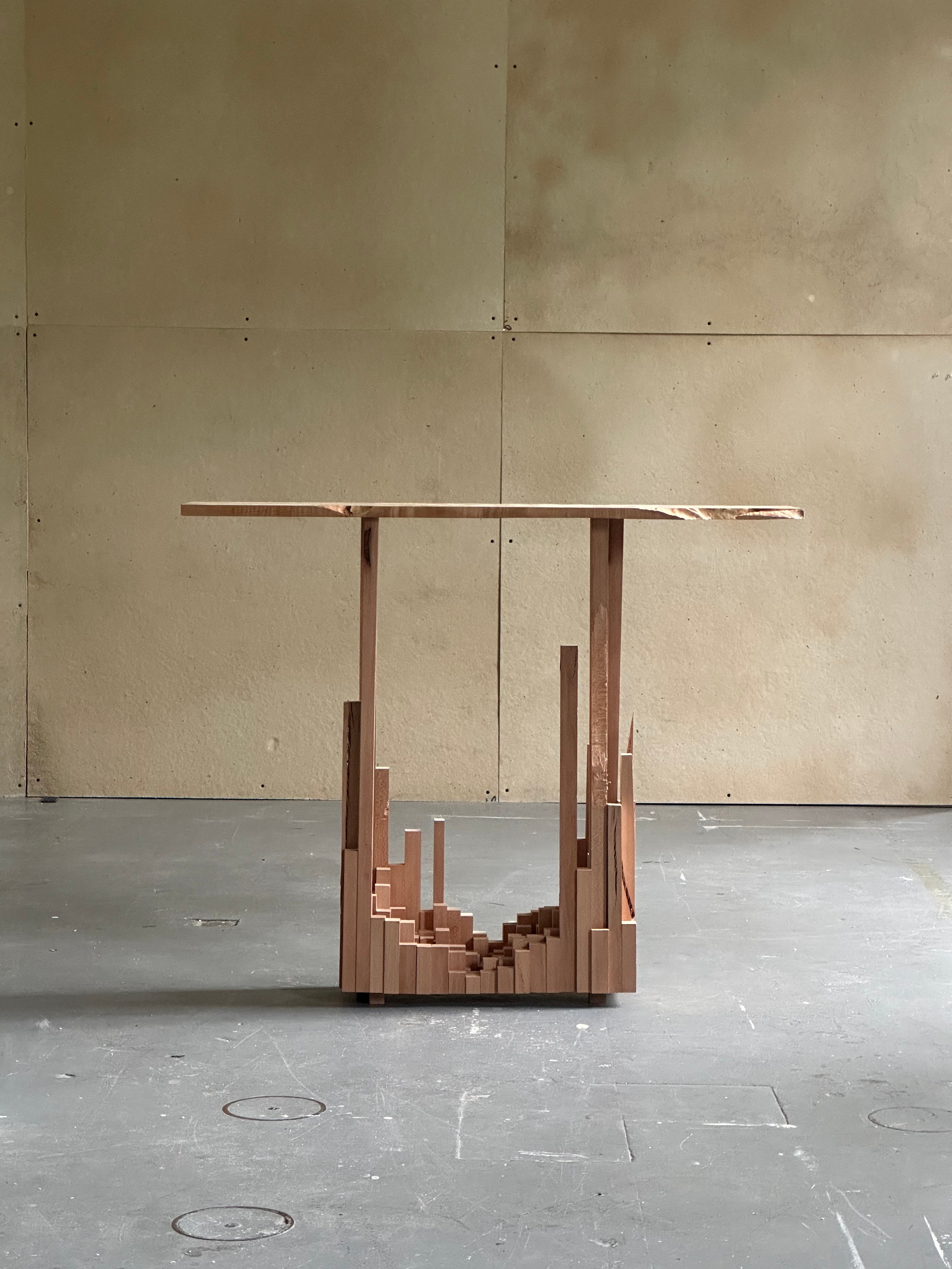 Minimalist wooden side table with a thin rectangular top supported by geometric vertical elements rising from clustered blocks. Warm wood tones create an architectural, skyline-like composition.
