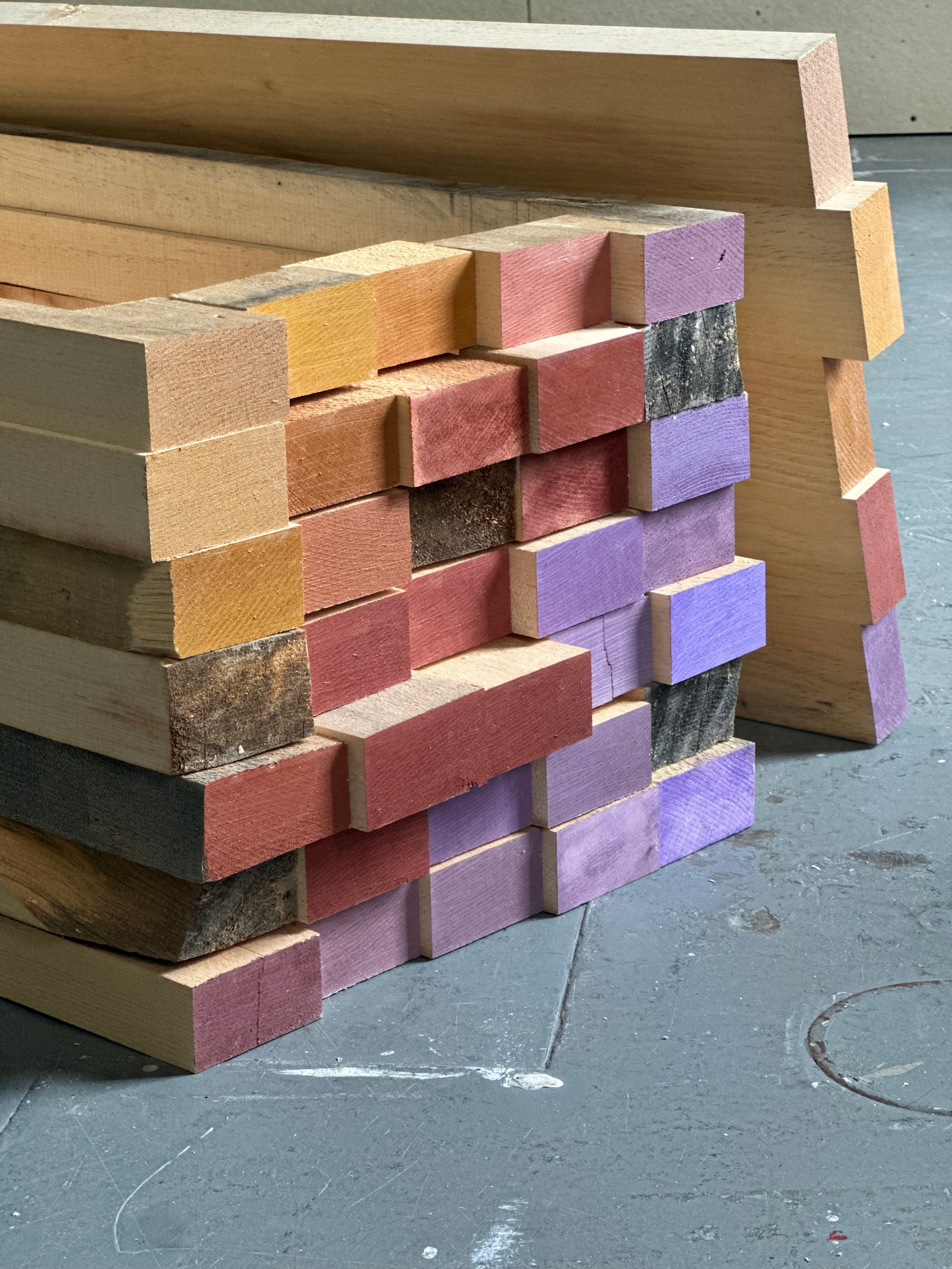 Stacked wooden blocks form a geometric table base with exposed end grain, varied wood tones, and painted surfaces. The layered construction emphasizes repetition, material contrast, improvisation, and the tension between rough construction and careful composition.