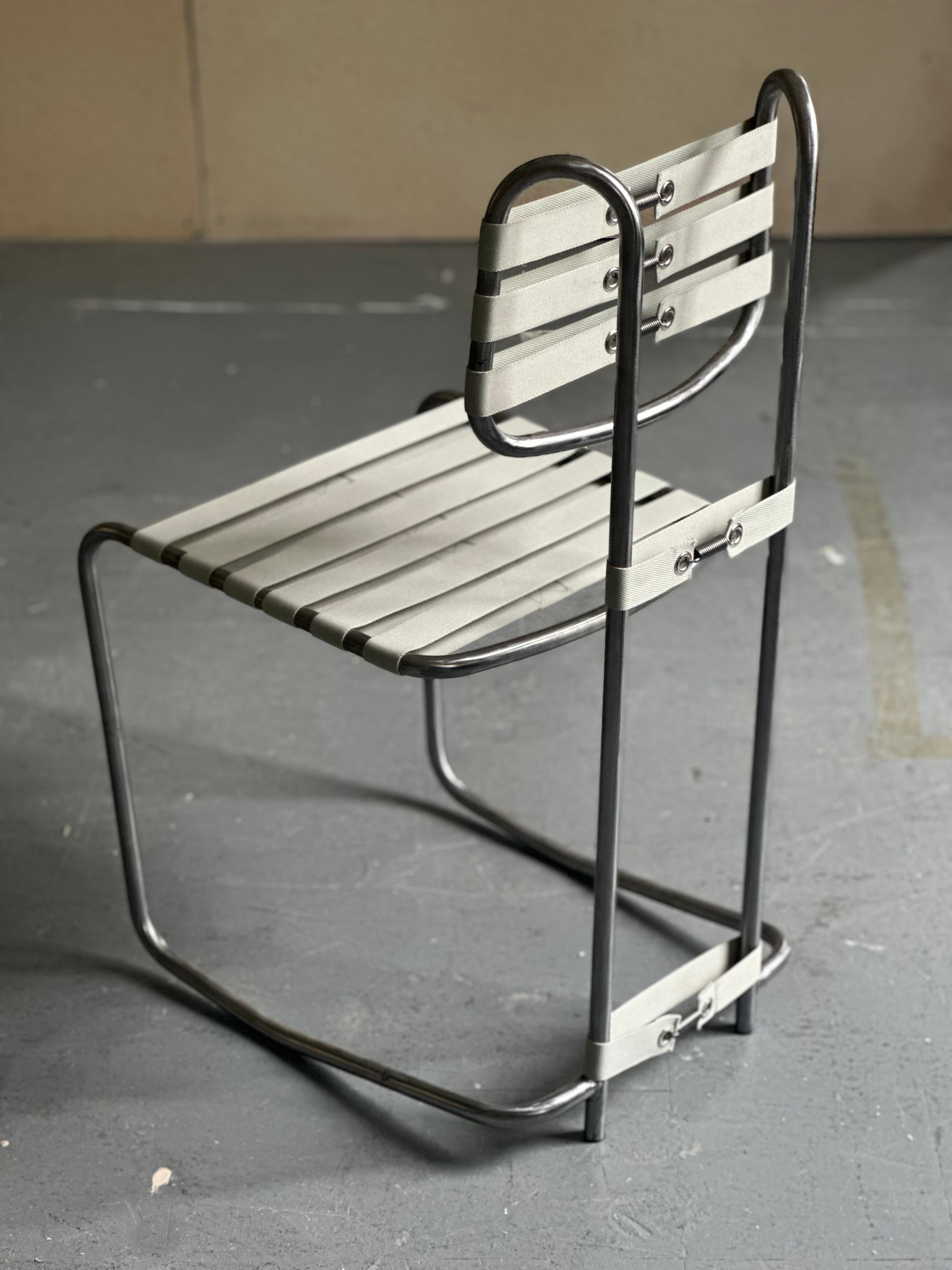 Cantilevered steel tube chair with tensioned nylon webbing seat and backrest. The continuous black frame contrasts with pale strapping, emphasizing flexibility, visible structure, active sitting, and the relationship between tension, support, and movement.