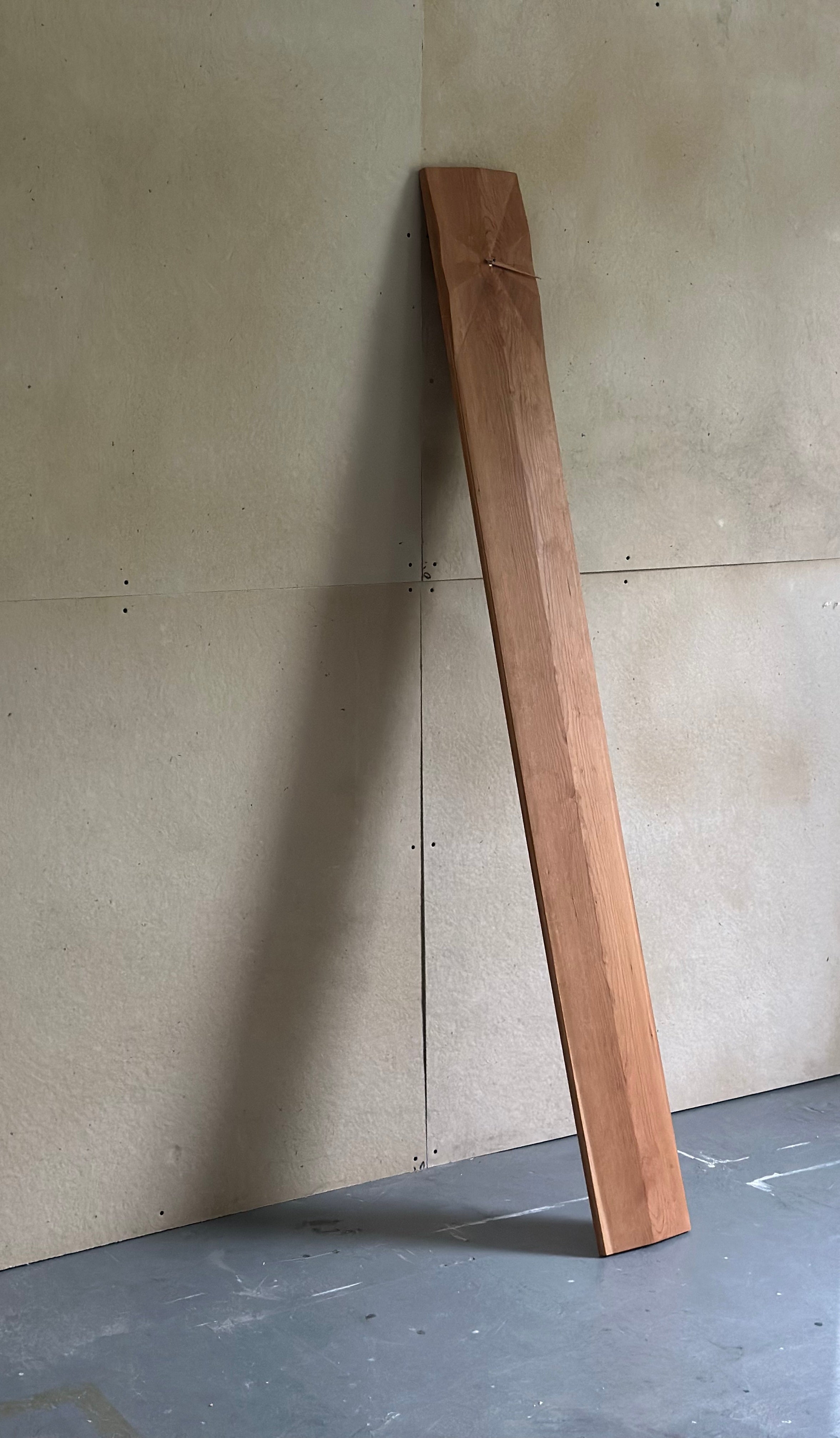 Minimal modernist cherry grandfather clock leaning casually against the wall, featuring a single hour hand and visible grain. The face is carved with a 12 radial facets.