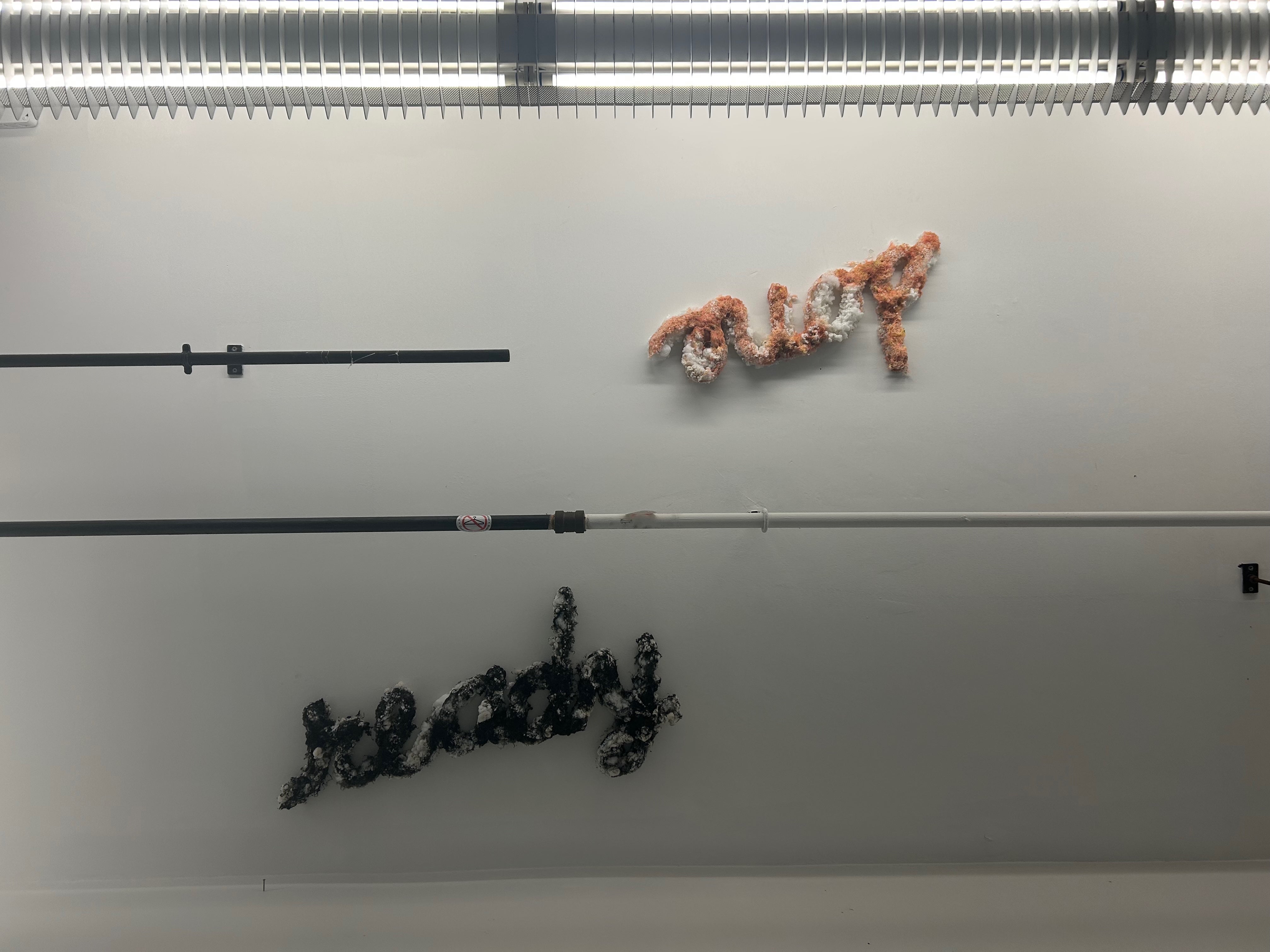 Ceiling shot, split by sprinkler piping and bordered by a florescent light. Cursive lettering-made from melted plastic shavings, stuffed animal fluff and pellets-is attached to the ceiling.