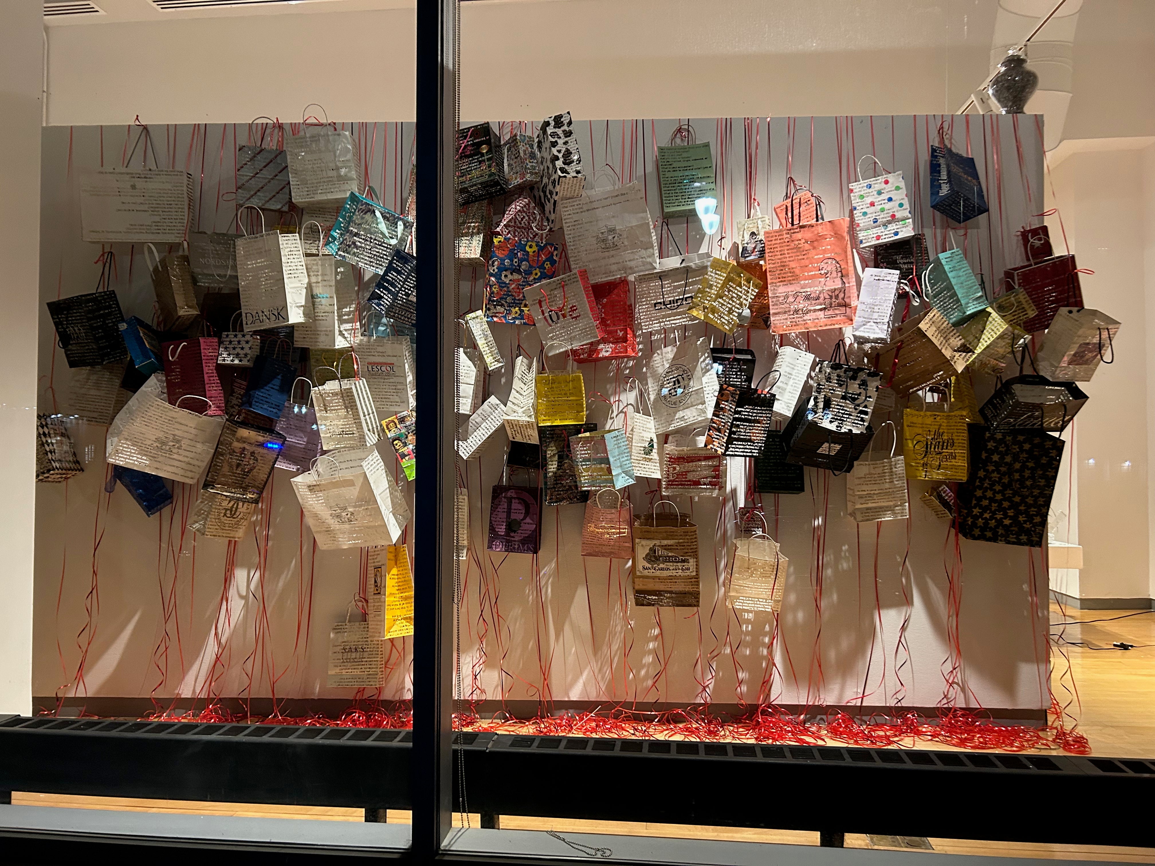 Window view of paper gift bags laser cut and hanging over a dividing wall. Variety of colors and sizes hanging from red party ribbon which spools and the floor beneath. Light casts shadows that show the cutouts.
