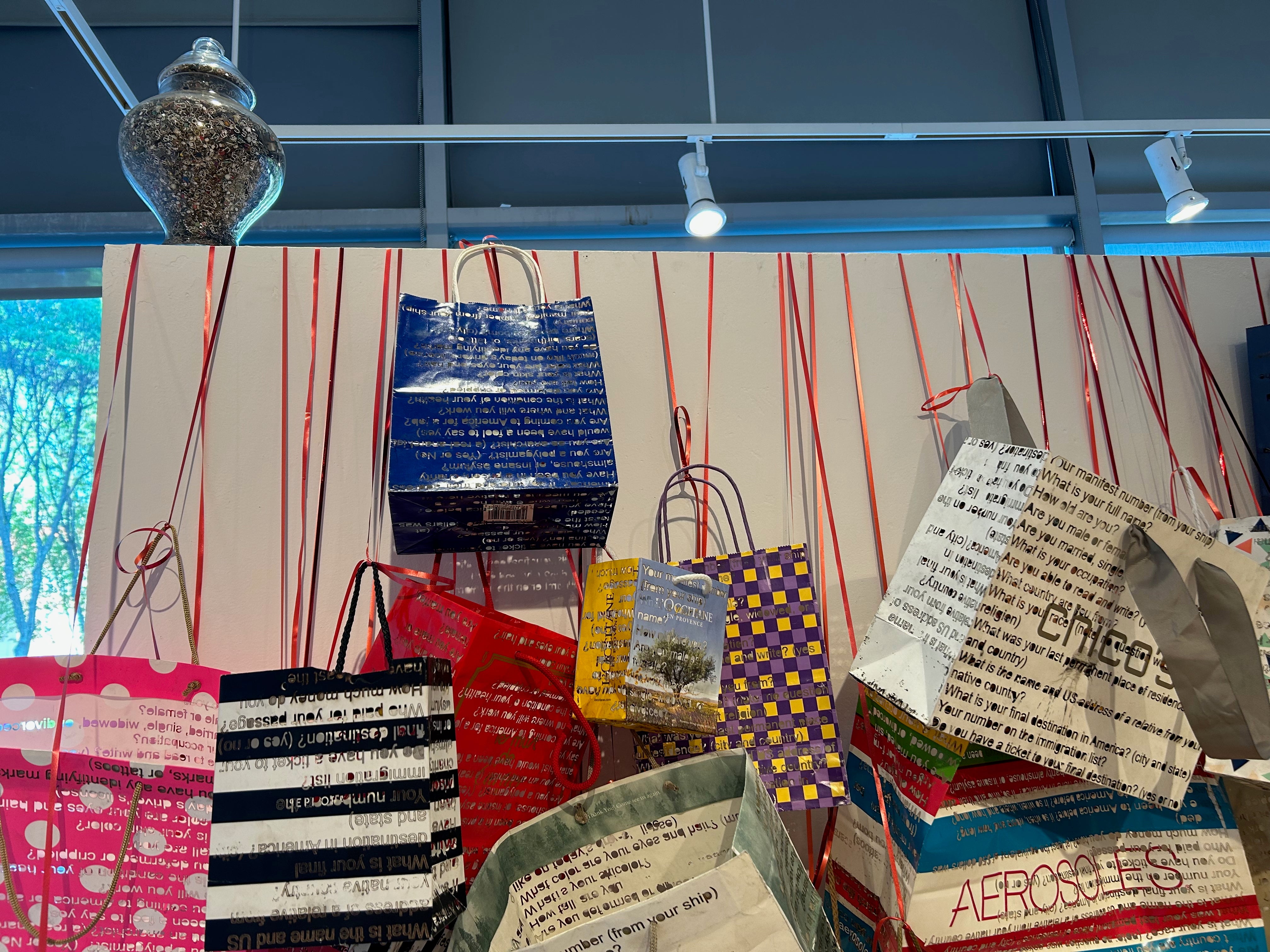 Close up of a few bags towards the top of the wall. Lettering is readable as the series of questions asked at Ellis Island. Glimpse of how the light casts through the cutouts. Letters in the