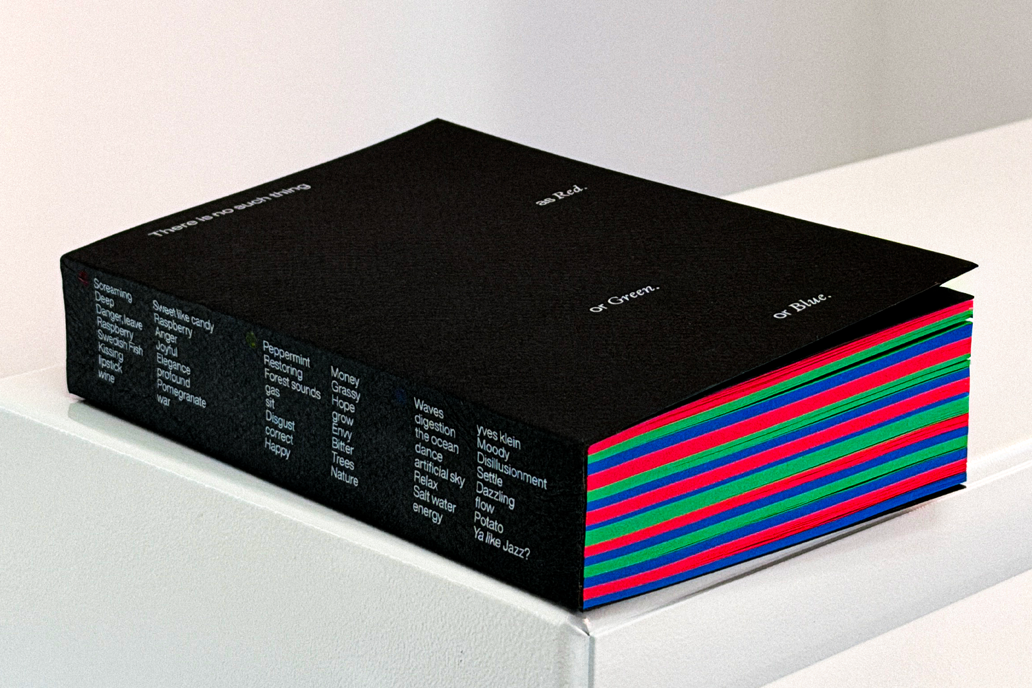 A 600-page book titled "There is no such thing as Red. or Green. or Blue." rests on a white pedestal. The spine lists participant survey responses. Page edges show alternating red, green, and blue stripes.