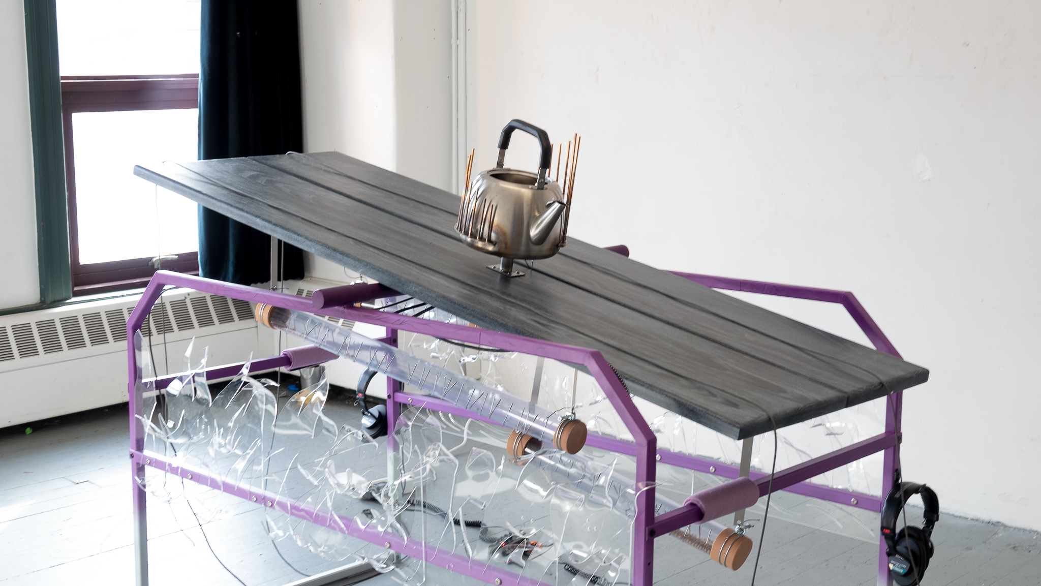 Purple steel table with kettle on wooden platform, ropes at both ends, clear acrylic sides showing rain sticks beneath, headphones attached.