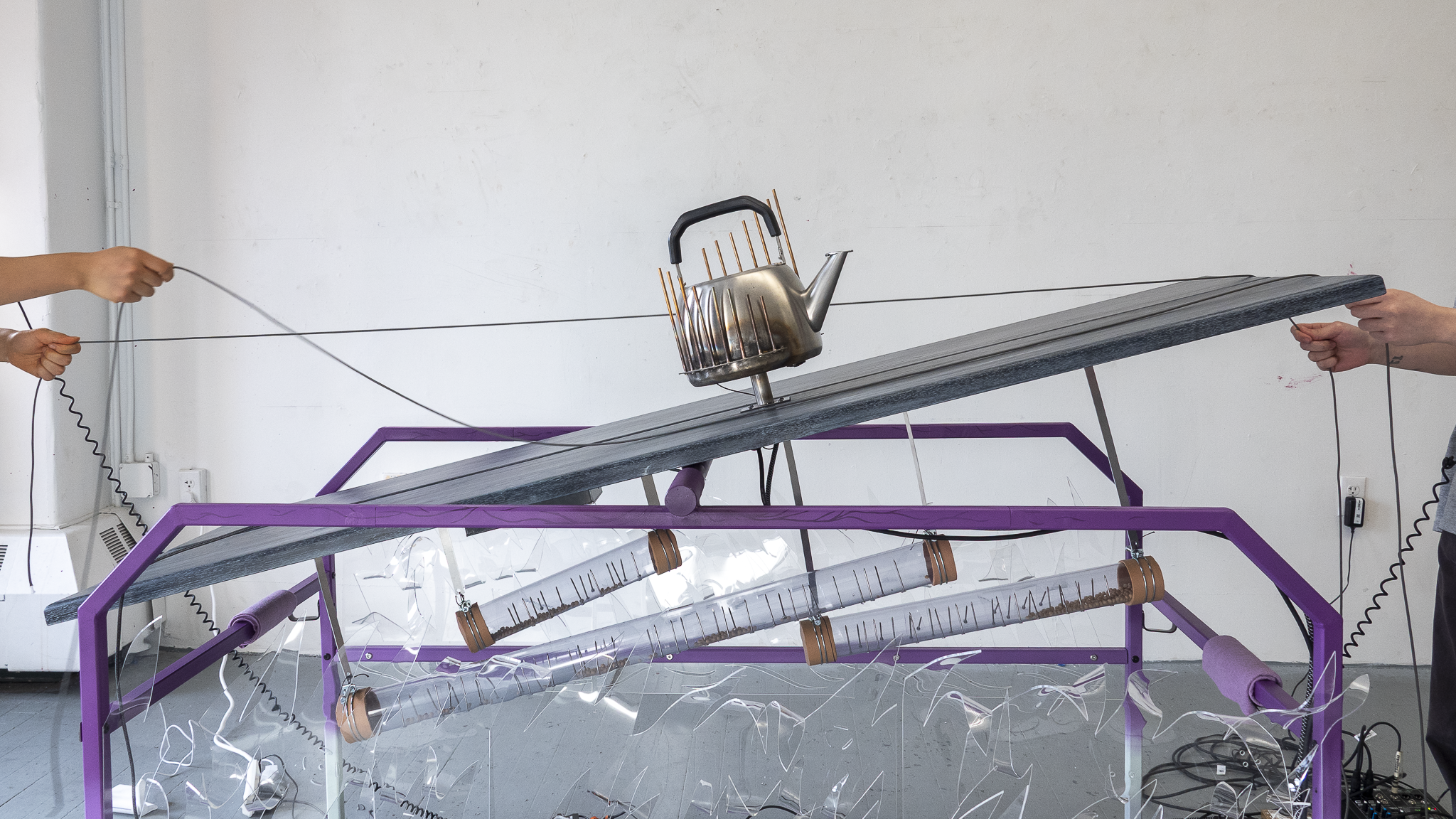 Two people pull ropes at opposite ends of a purple table, tilting a wooden platform with a waterphone-like kettle on top.