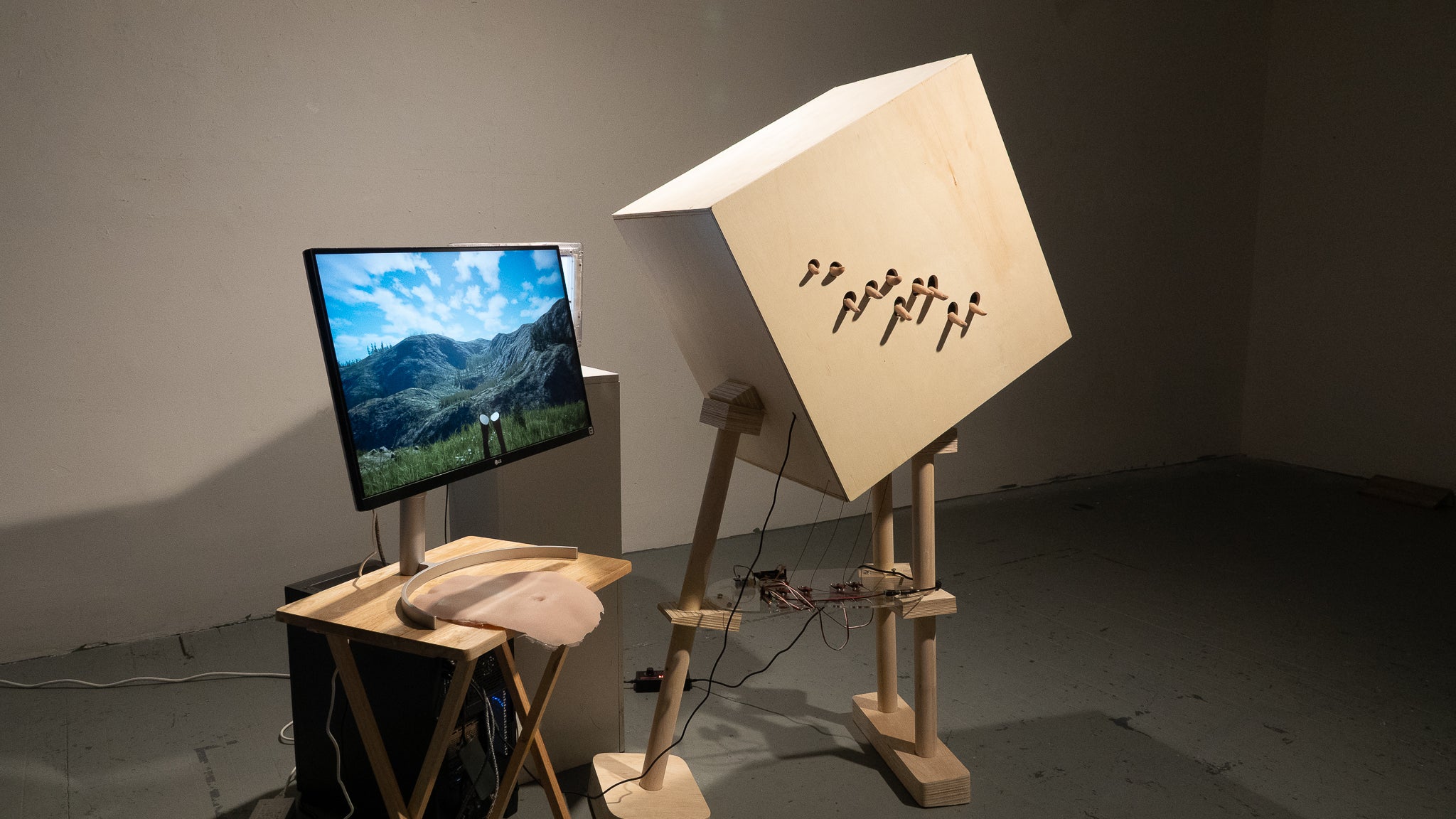 Installation with monitor showing digital landscape, silicone interface, and wooden structure with protruding fingers.