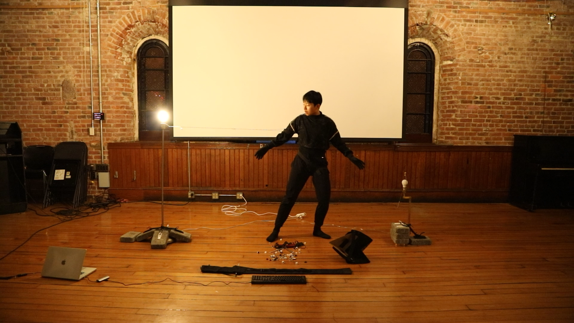 Performer in black stands among cables, props, and projection equipment in a brick-walled room.