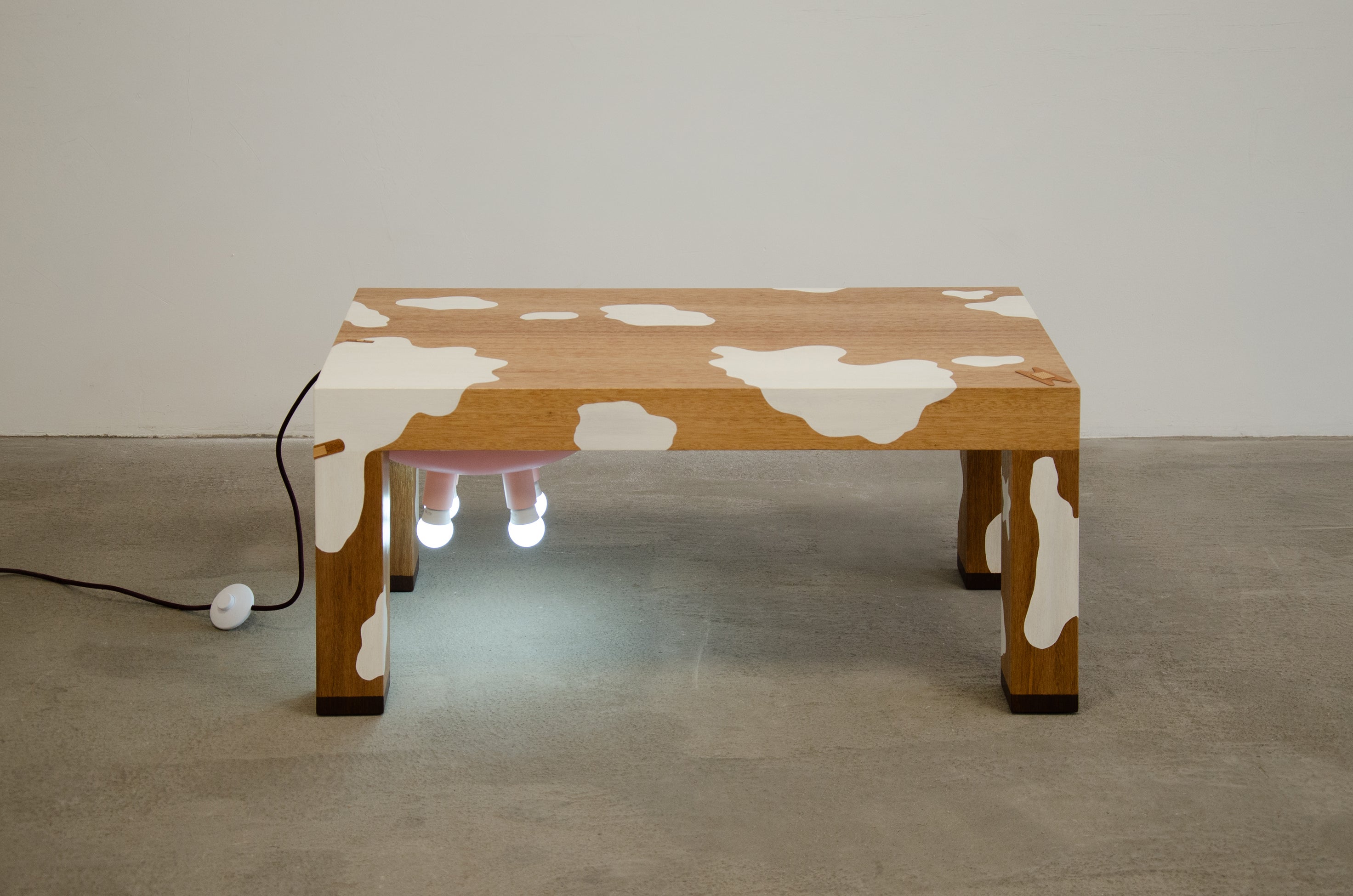 A cow-inspired wooden coffee table with a brown body, a pale pink belly, four pink light bulbs underneath like udders, and a cord extending from the back like a tail.