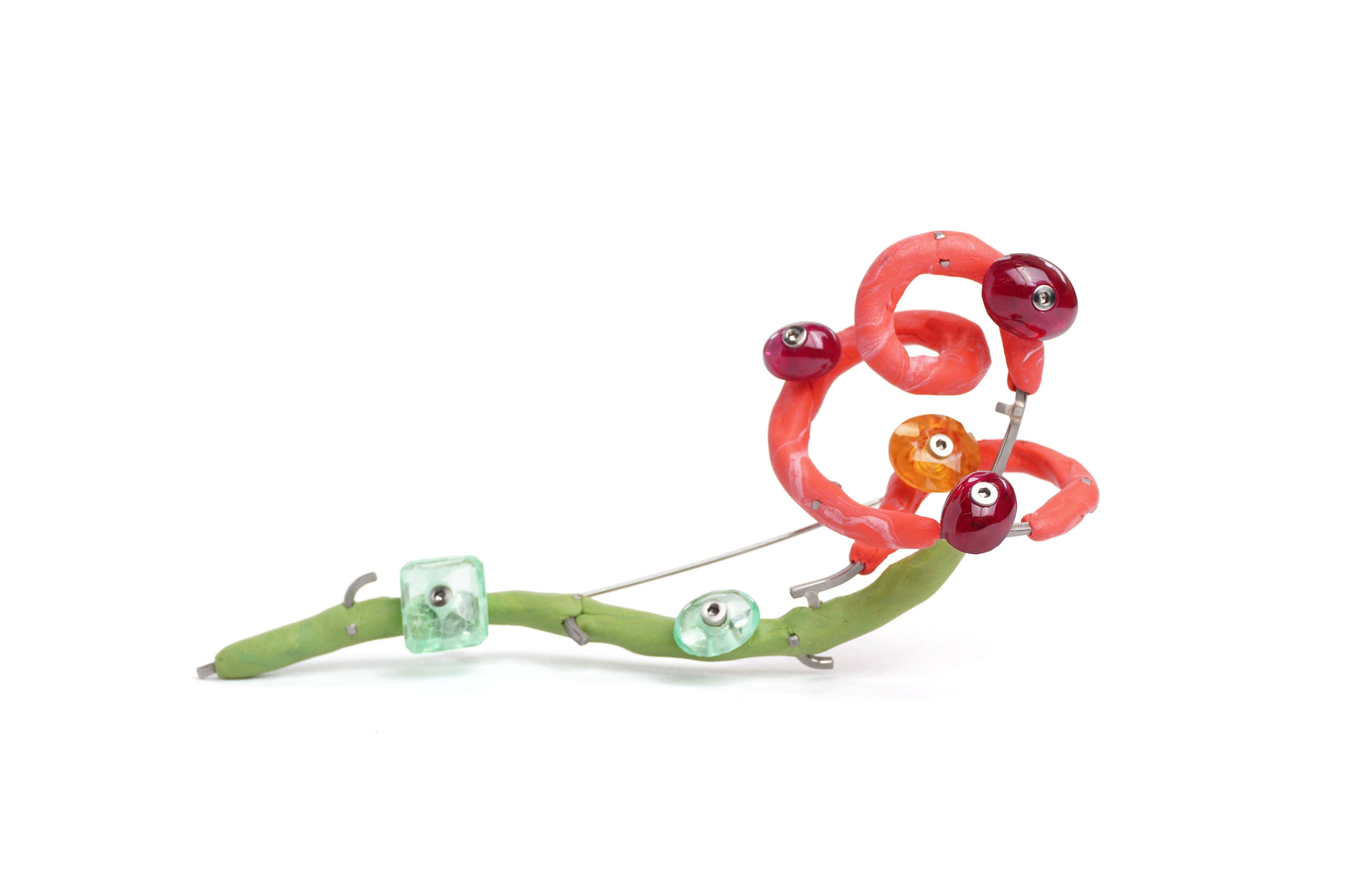 A brooch loosely in the shape of a rose, made up of marbled pink and green silicone, and metal wire. Rubies and sapphires are bolted along its length with screws. On a white background.