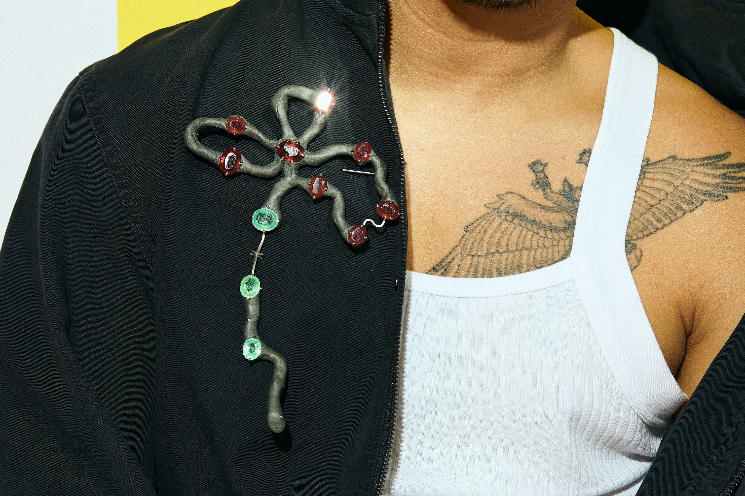 A brooch loosely in the shape of a flower with three large petals, made up of grey epoxy and metal wire. Rubies and sapphires are set in prong settings along its length. Worn by a man with a vest, closely cropped to his chest.