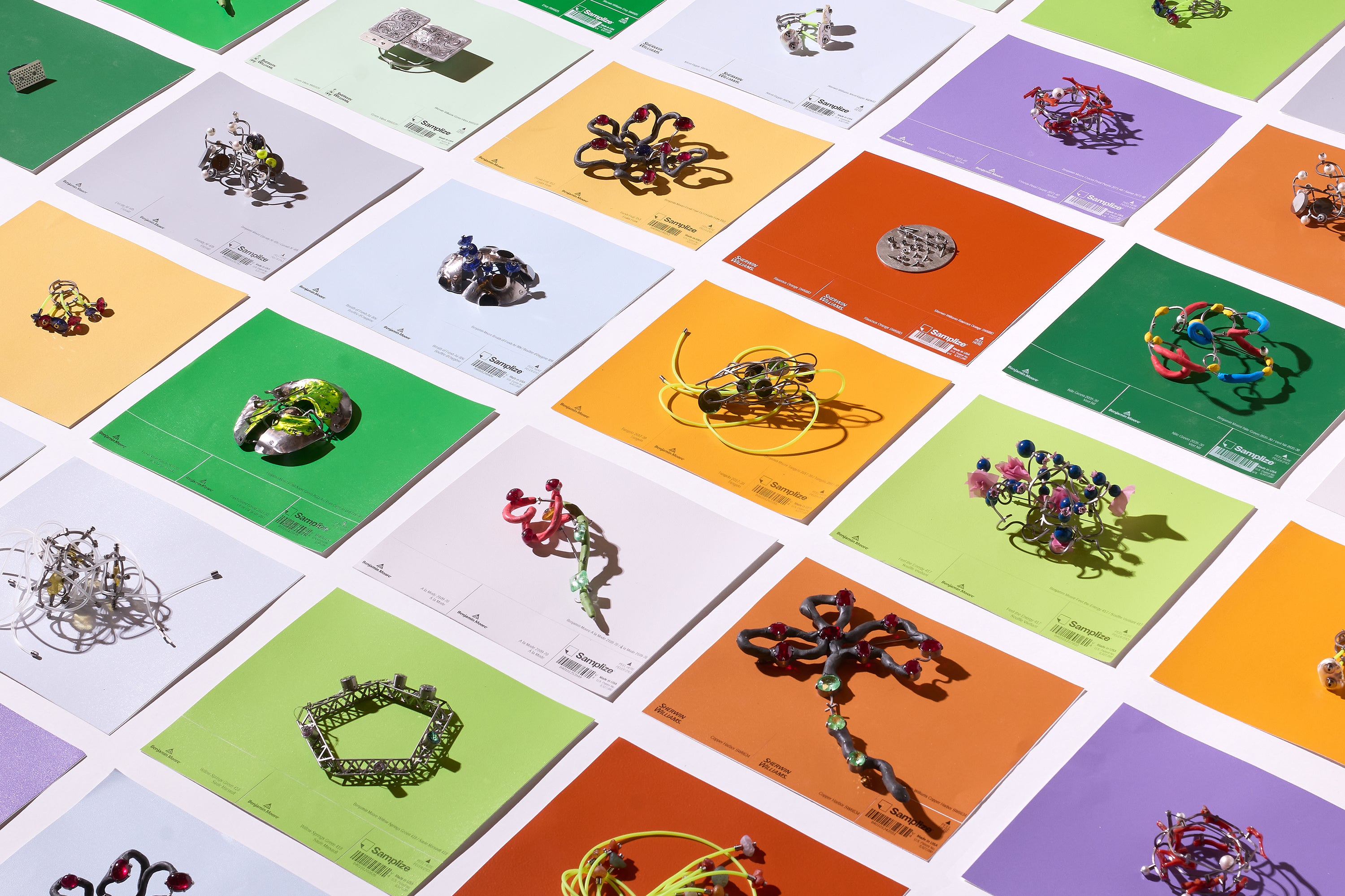 An isometric perspective of approximately 23 rainbow coloured tiles laid out on a white floor. There is a piece of jewellery on each tile, receding into the background. The jewels include rings, pendants, brooches, and bracelets.