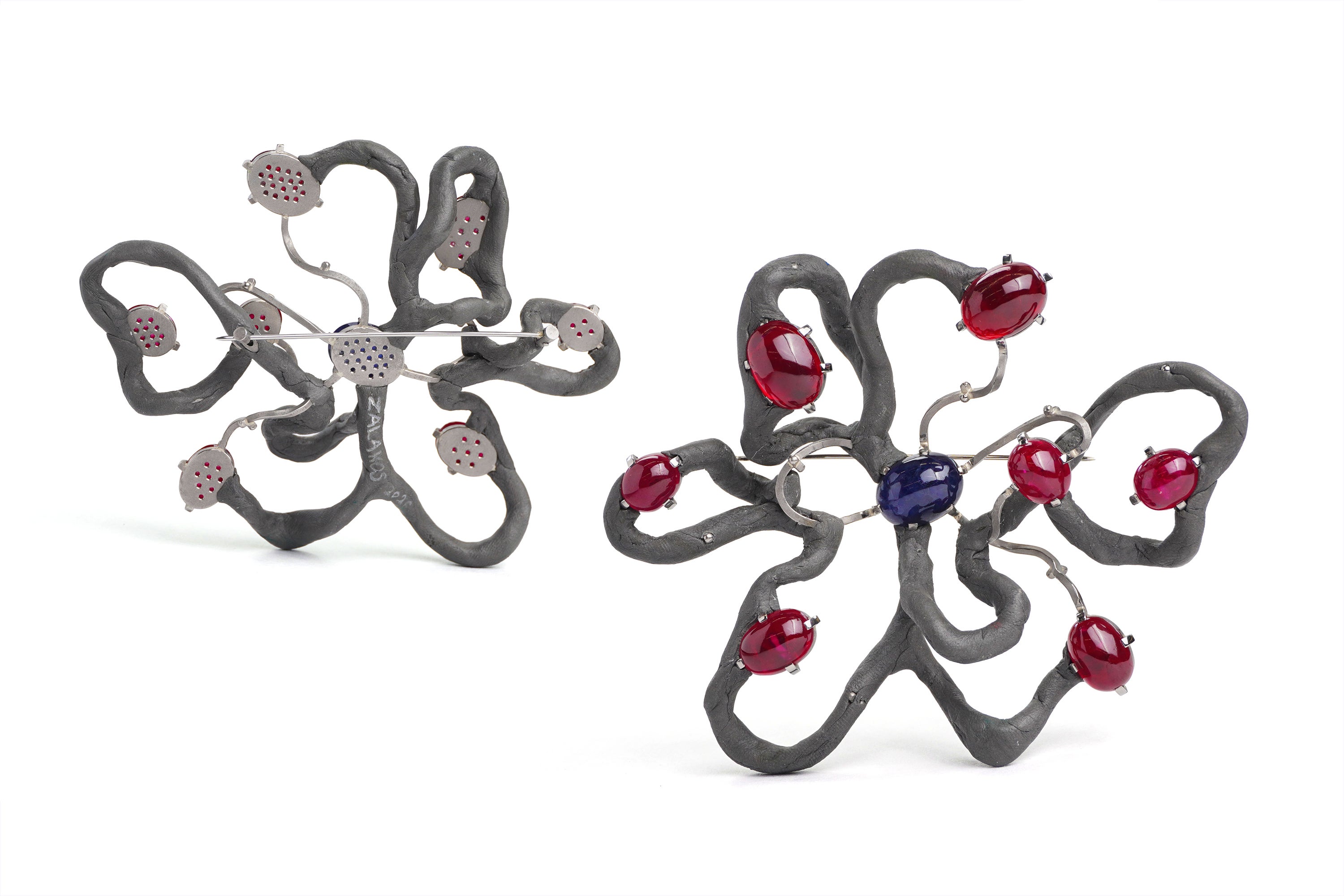 A brooch loosely in the shape of a flower with seven large petals, made up of grey epoxy and metal wire. Rubies and sapphires are set in prong settings along its length. The brooch is pictured