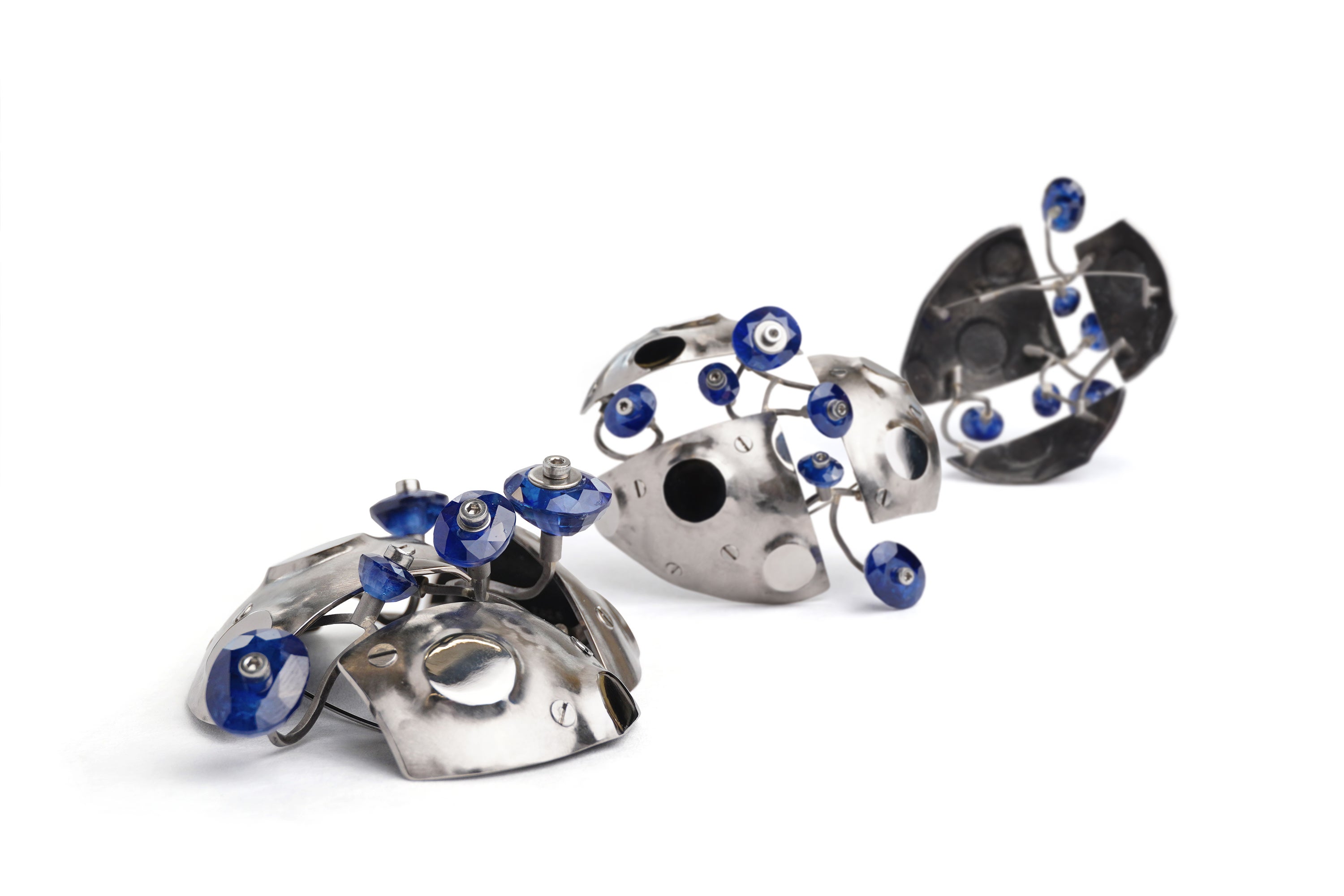 A brooch made from three rounded grey metal panels that are bolted together with gaps between them. Small circular mirror panels are raised from its surface with polished faces. Short lengths of twisted square wire branch