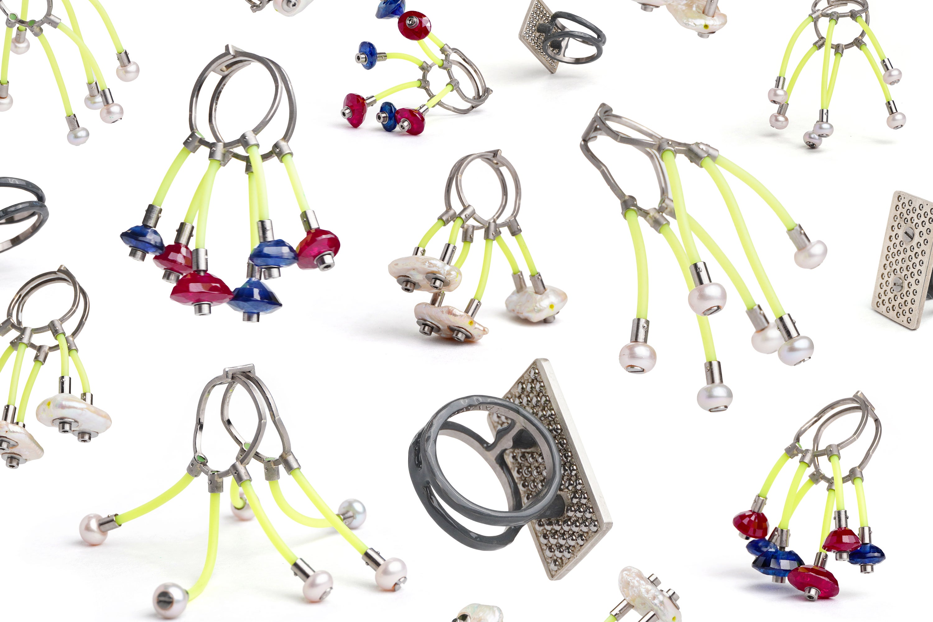 A composite image of four rings pictured from sixteen different angles on a white background. The rings have pearls, sapphires, or silver panels bolted onto bright yellow lengths of rubber that are connected to the ring shanks made from loops of square wire.