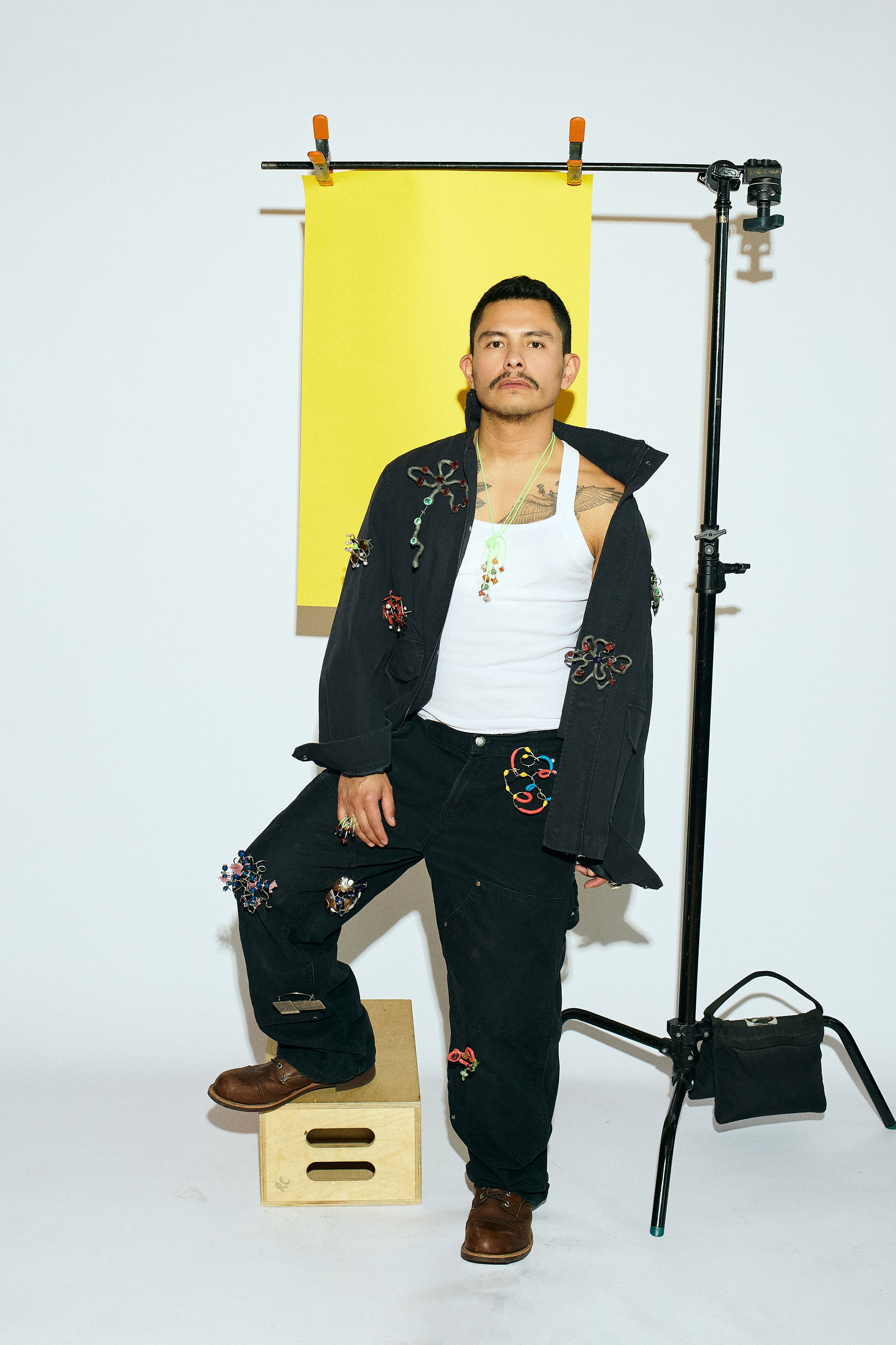 A man in workwear standing in a photo studio on a white background. A tripod beside him holds a sheet of yellow paper behind his head. He has twelve brooches pinned to his clothes and wears rings and pendants.