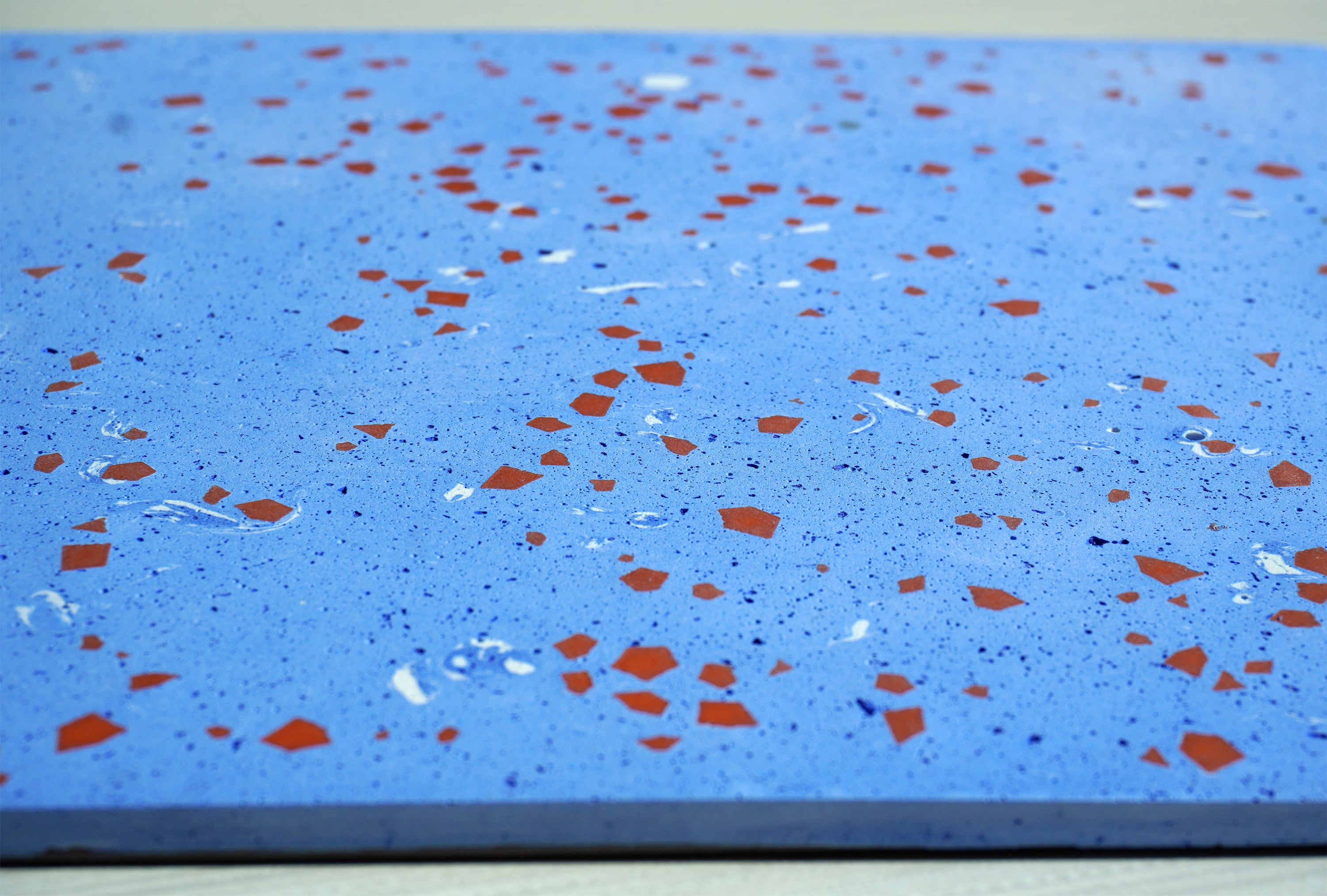 Side-angle close-up of a pale blue terrazzo tile showing depth and terracotta geometric fragments embedded in the surface.