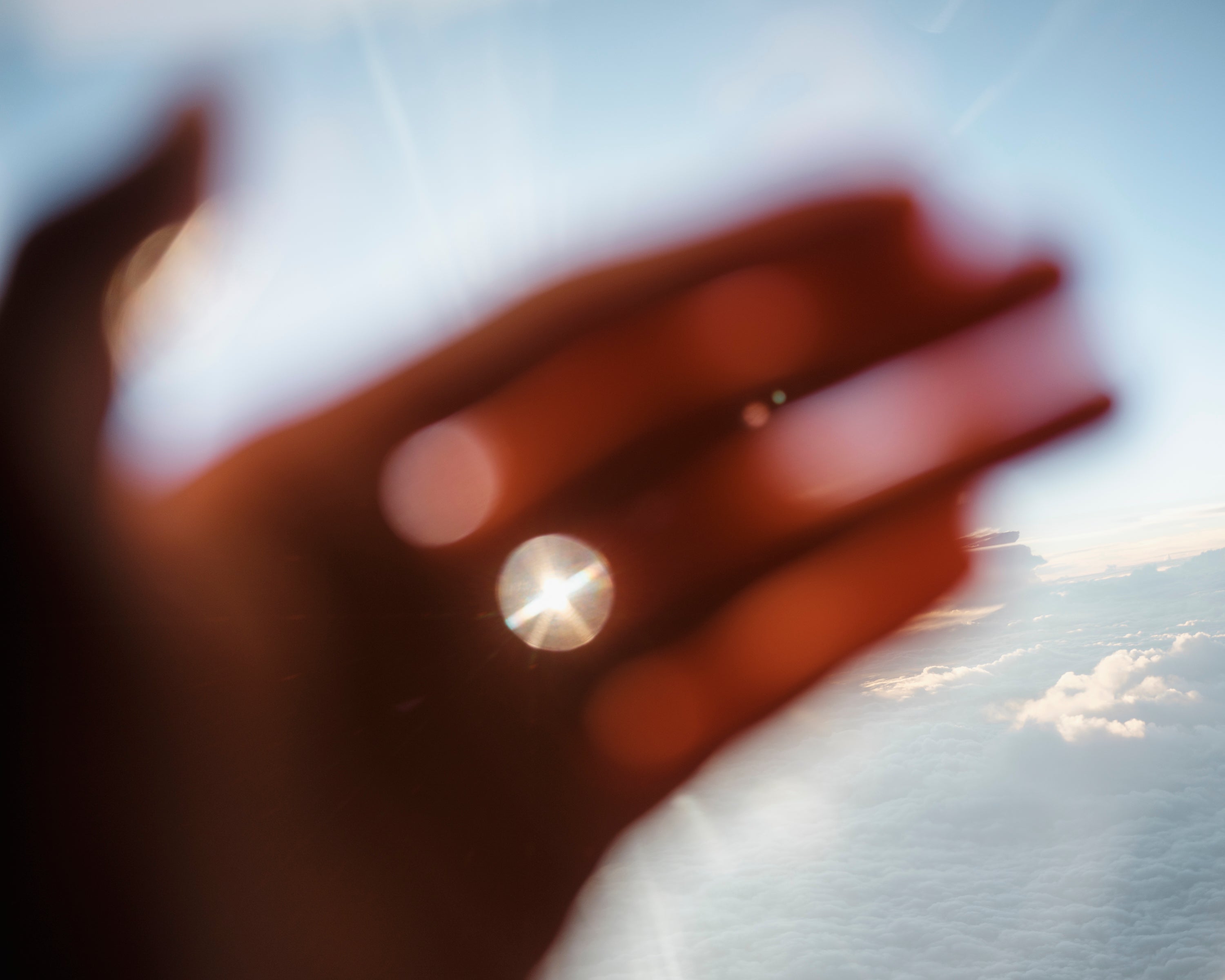 Color photograph of the palm of a hand glowing in orange, held in front of a bright blue sky, with the sun peeking through the gap between the fingers.
