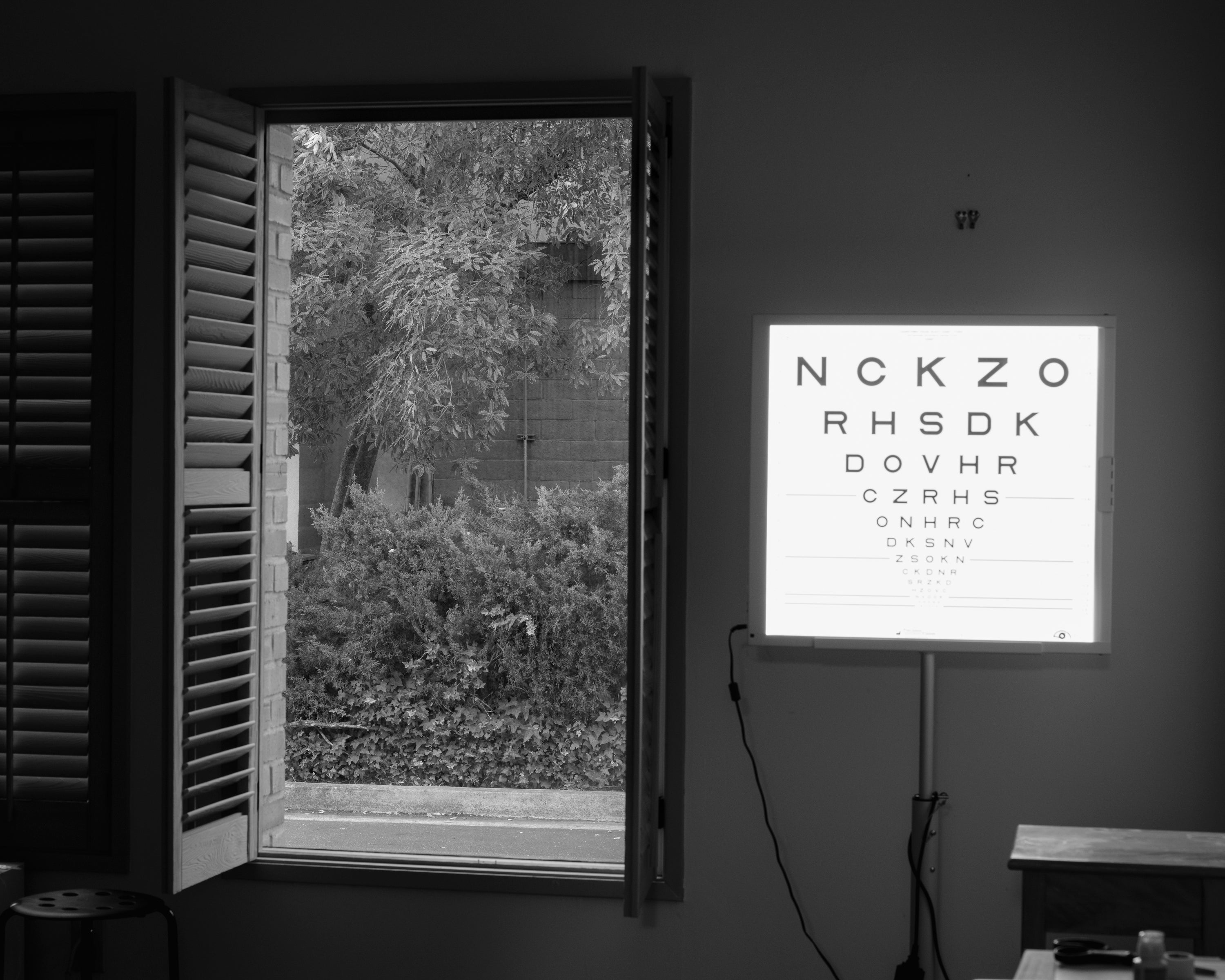 Black and white photograph of inside the vision test room. A soft glowing vision test chart illuminates the right side of the room. To the left, a tall window with its wooden blinds open, shows trees and plants outside.