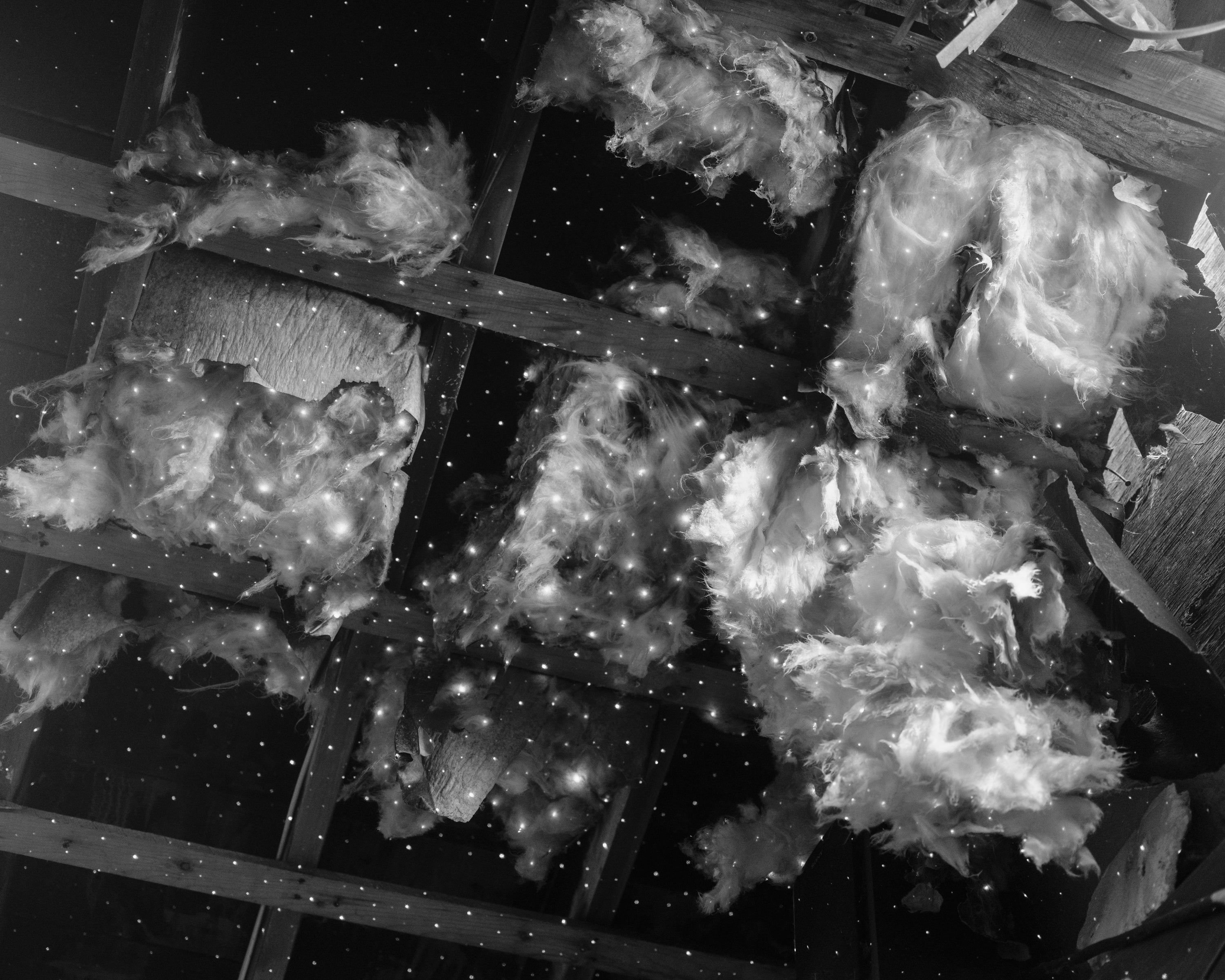 Black and white photograph of tiny white dots of light glimmering like stars, on a collapsed ceiling. Fiberglass insulation materials are falling out from the wooden frames of the ceiling.