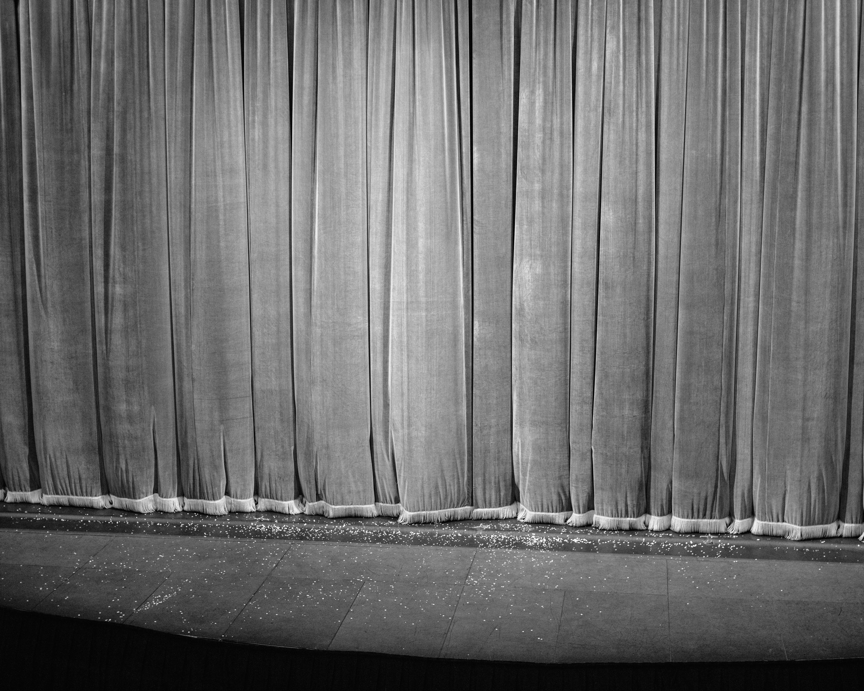 Black and white photograph of a thick velvet curtain, closed, with tassels dragging on the stage floor. Small white flakes are sprinkled on the floor.