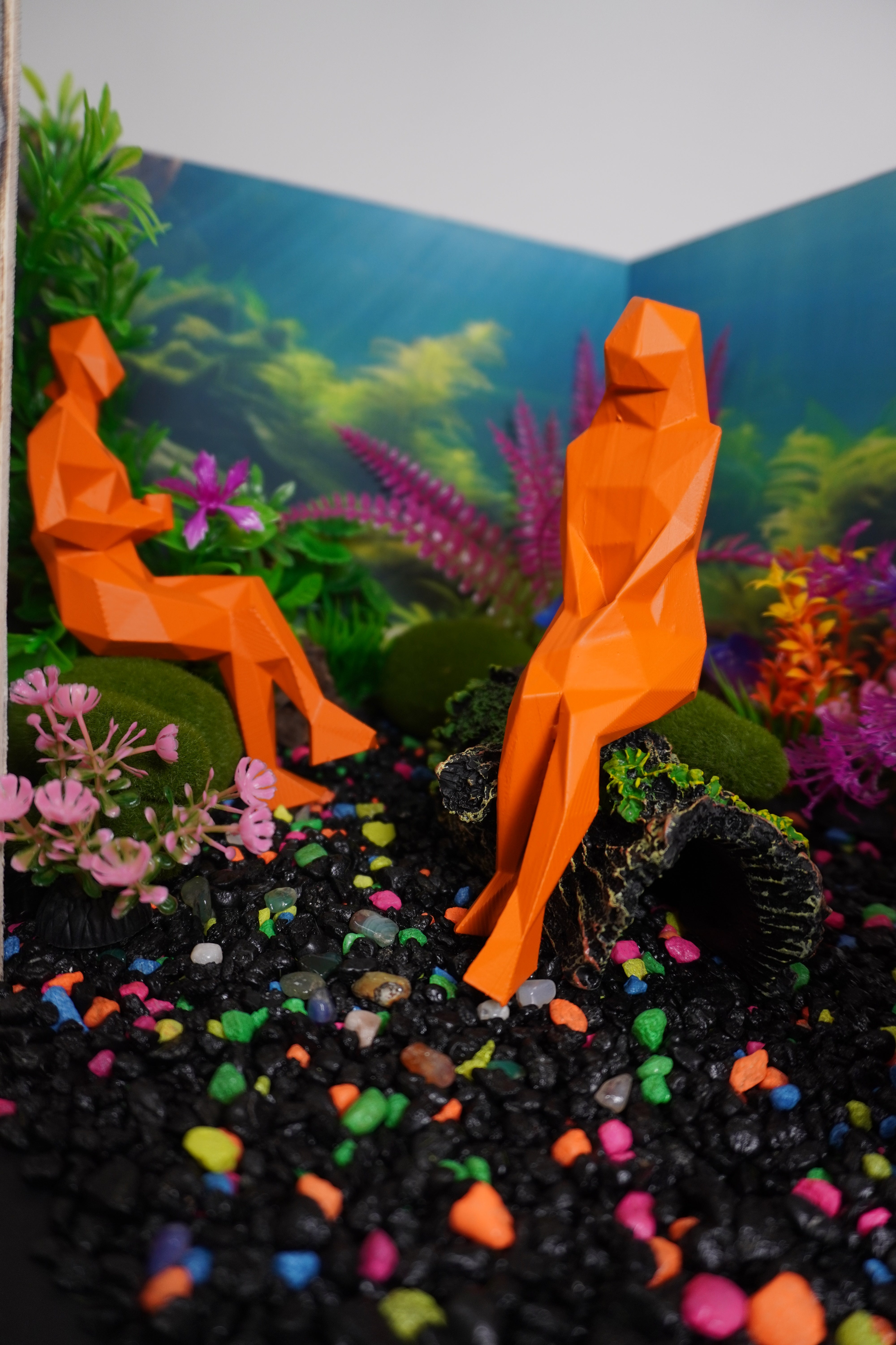 Photo of a model of a waiting room designed to look like an aquarium: black gravel with neon, multi-colored pebbles mixed in; "moss" covered "rocks" and a fake, "moss" covered "log" act as seating for orange painted, angular human figures. Colorful plastic plants line the perimeter of the room and the walls are covered in a wallpaper mural of an underwater image with seaweed and sun streaks.
