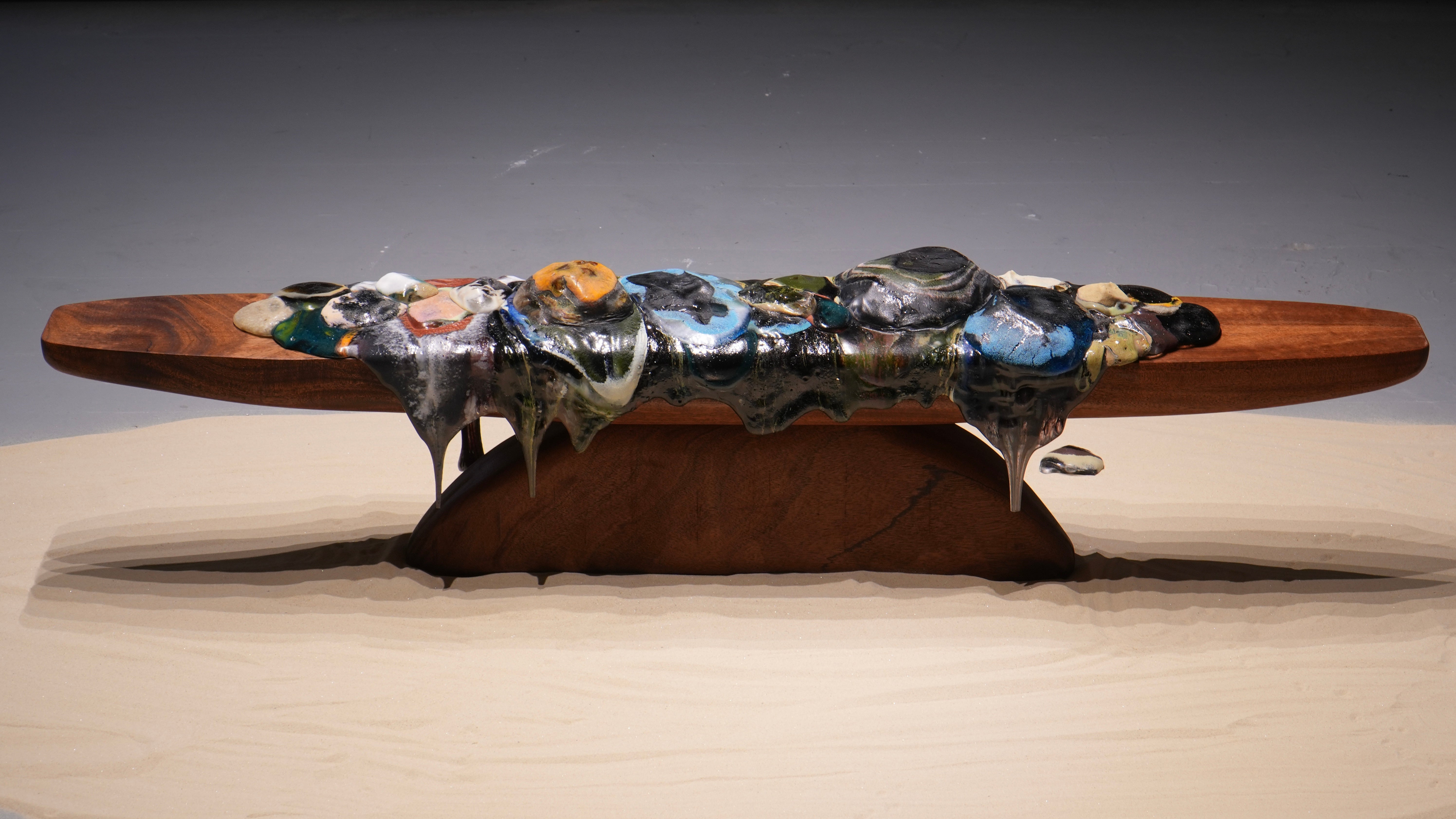 Molten glass forms in varied colors spread and drip across a long wooden platform, with softened edges and flowing material extending downward.