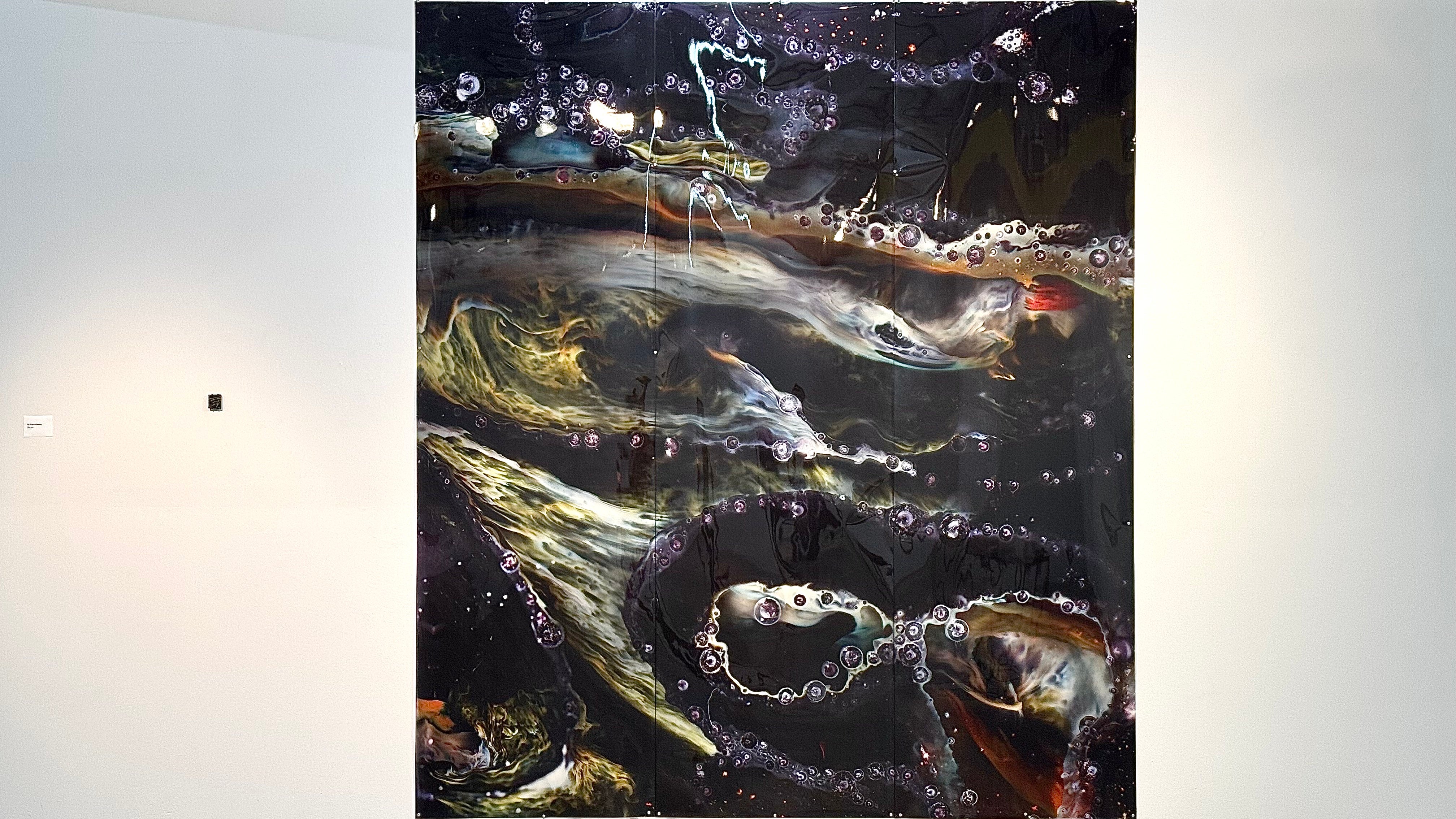 Large vertical print showing abstract patterns in dark tones with lighter flowing forms and bubble-like details, installed on a gallery wall along side with a small glass sample.