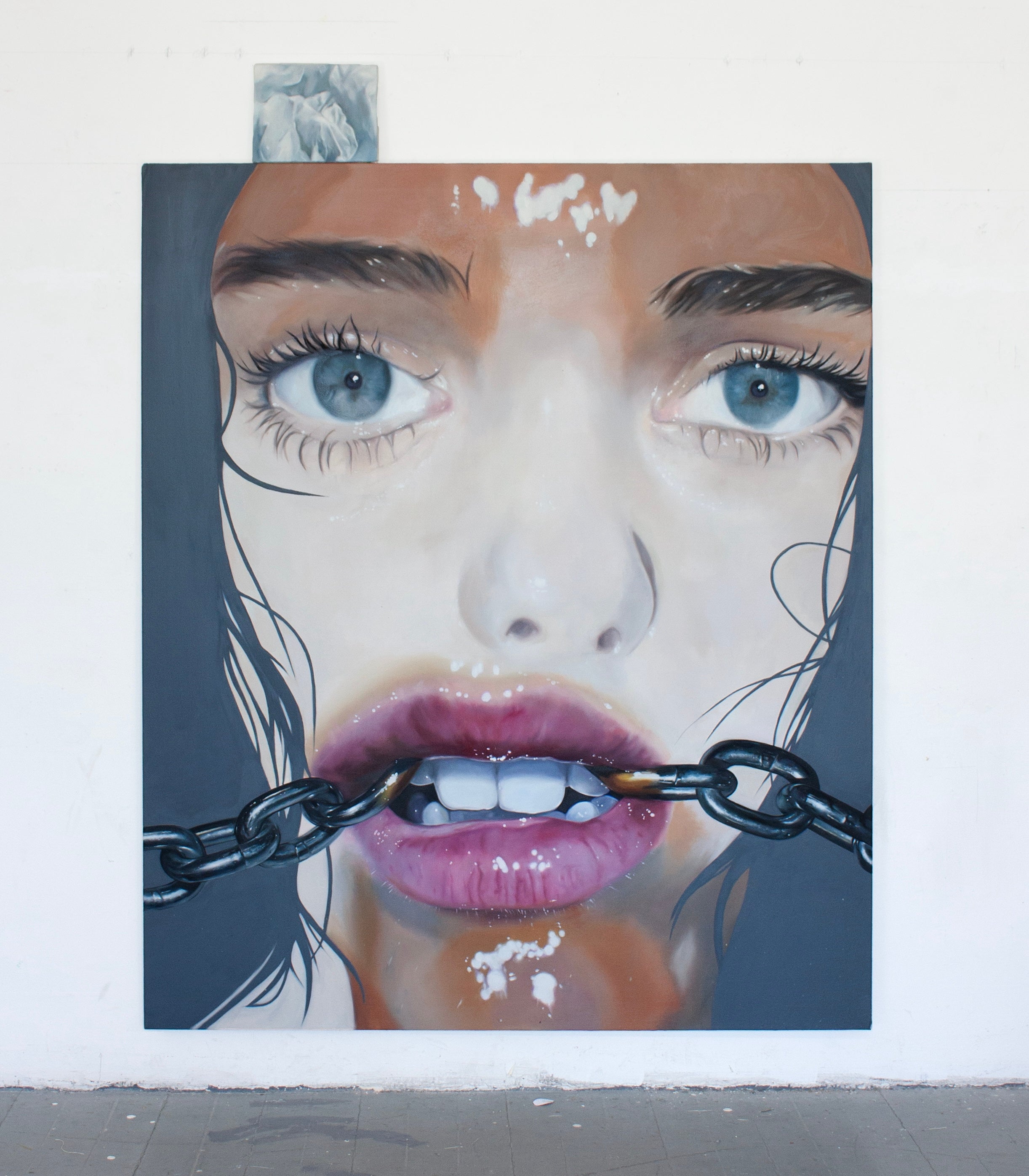 Large, close-up oil painted portrait of a woman's face. She has piercing blue eyes and parted lips with a chain extending out from both corners of her mouth.