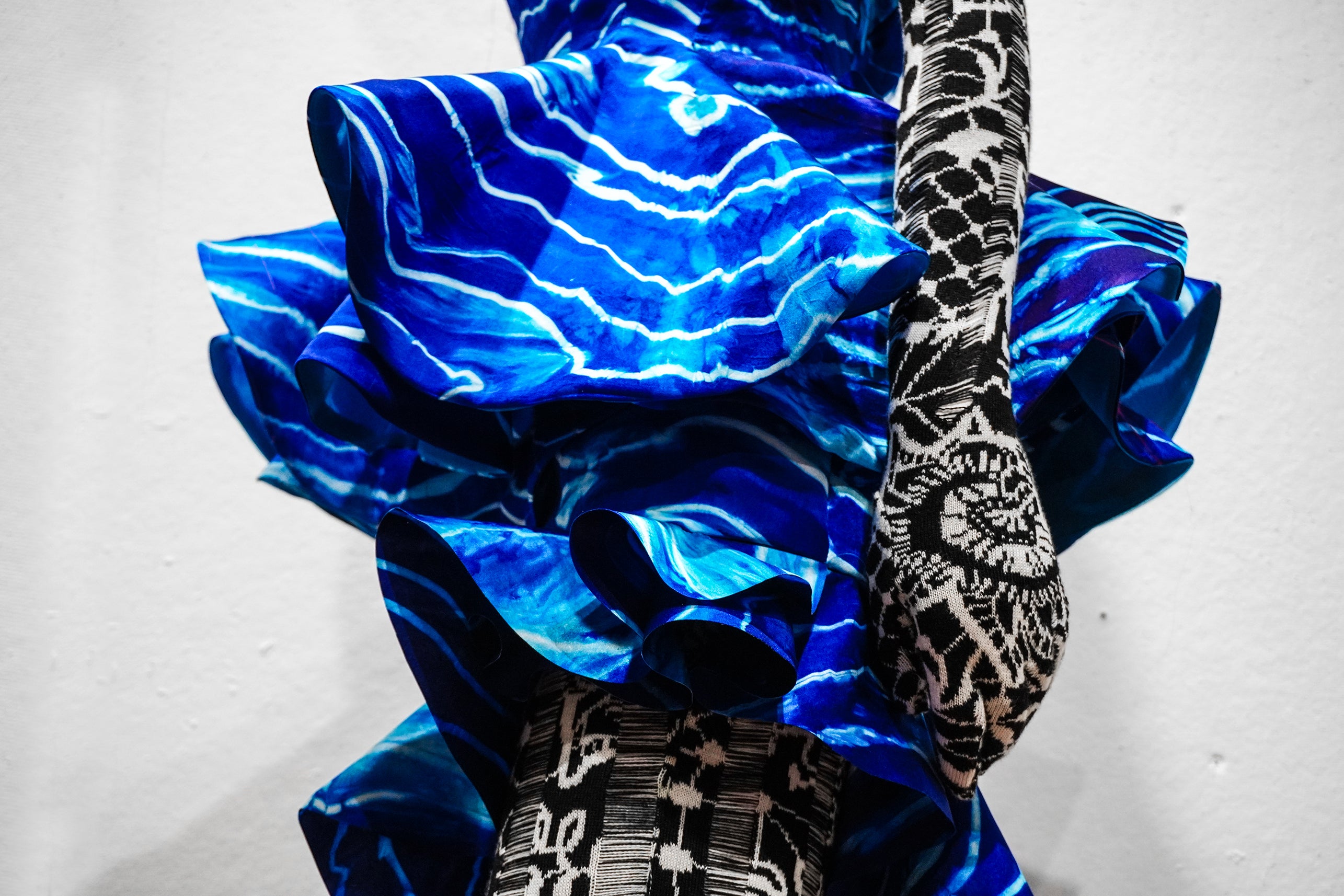 Close-up of sculptural blue silk ruffles with wave-like tie-dye patterns over black-and-white jacquard knit