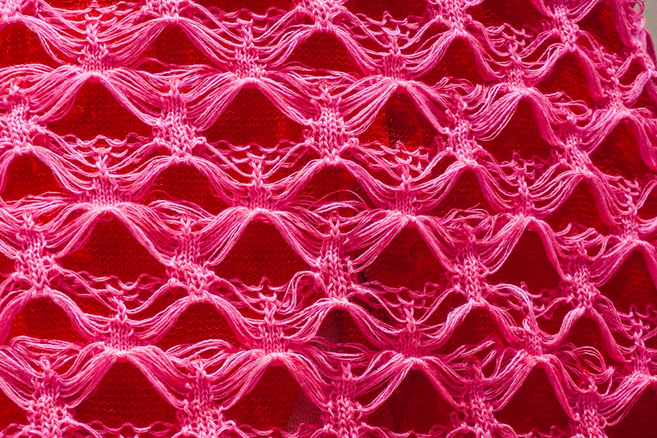 Close-up of bright pink knitted lace structure with looping openwork pattern over red textile background