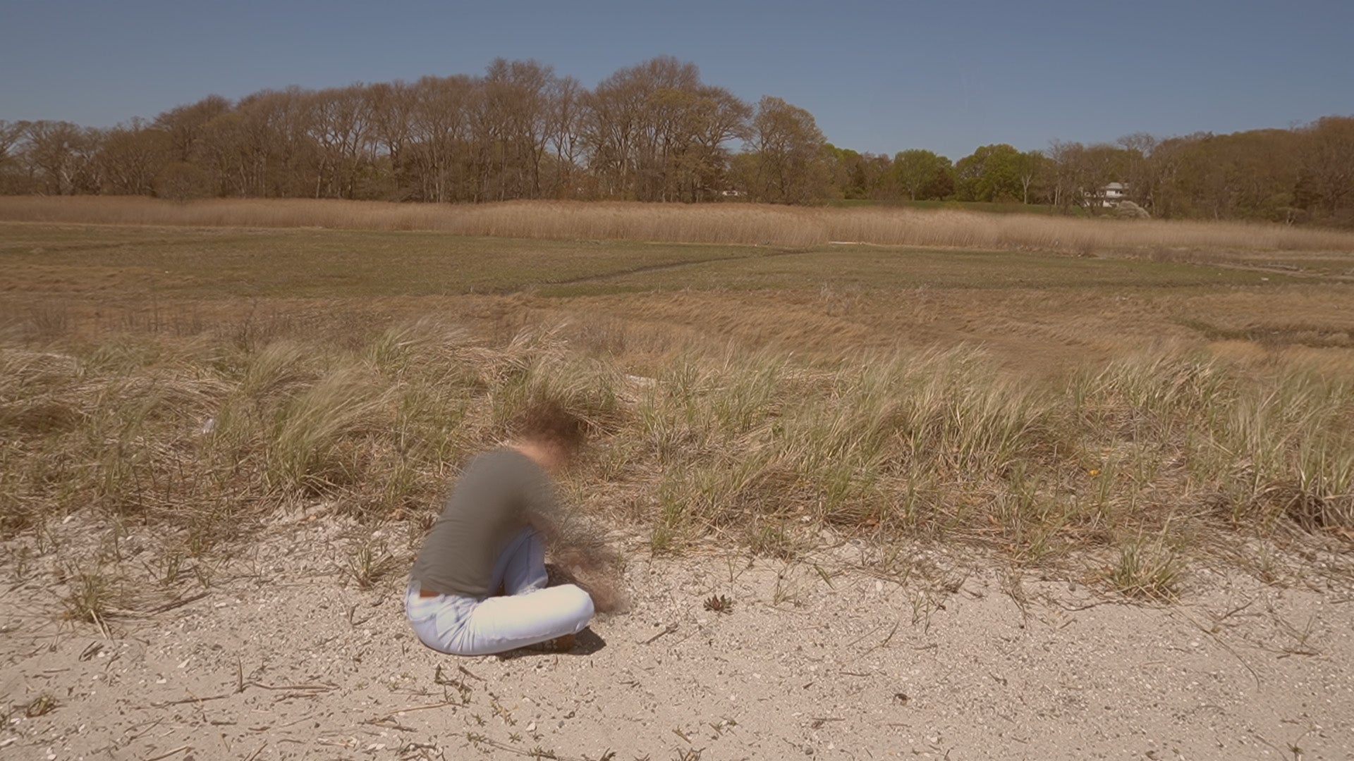 A person sits in a sandy dune landscape while making a bowl-shaped form in the ground. Marsh grasses and trees extend across the background beneath a clear sky.